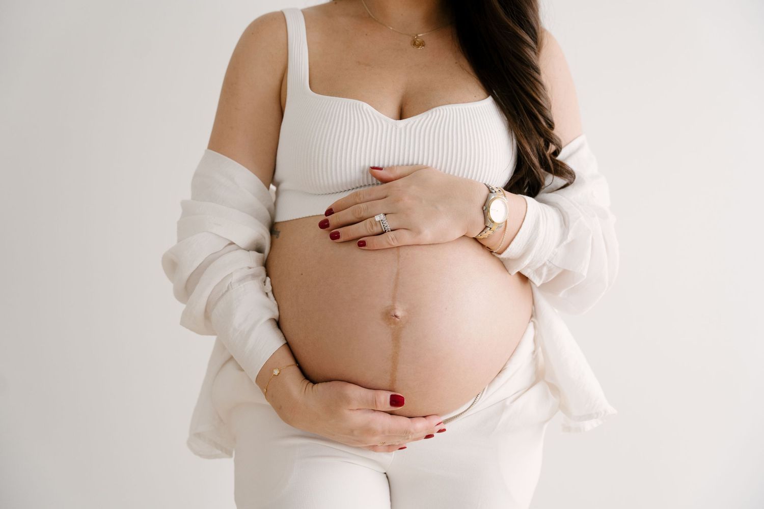 Close-up of pregnant woman in white crop top and cardigan gently holding baby bump with manicured hands.