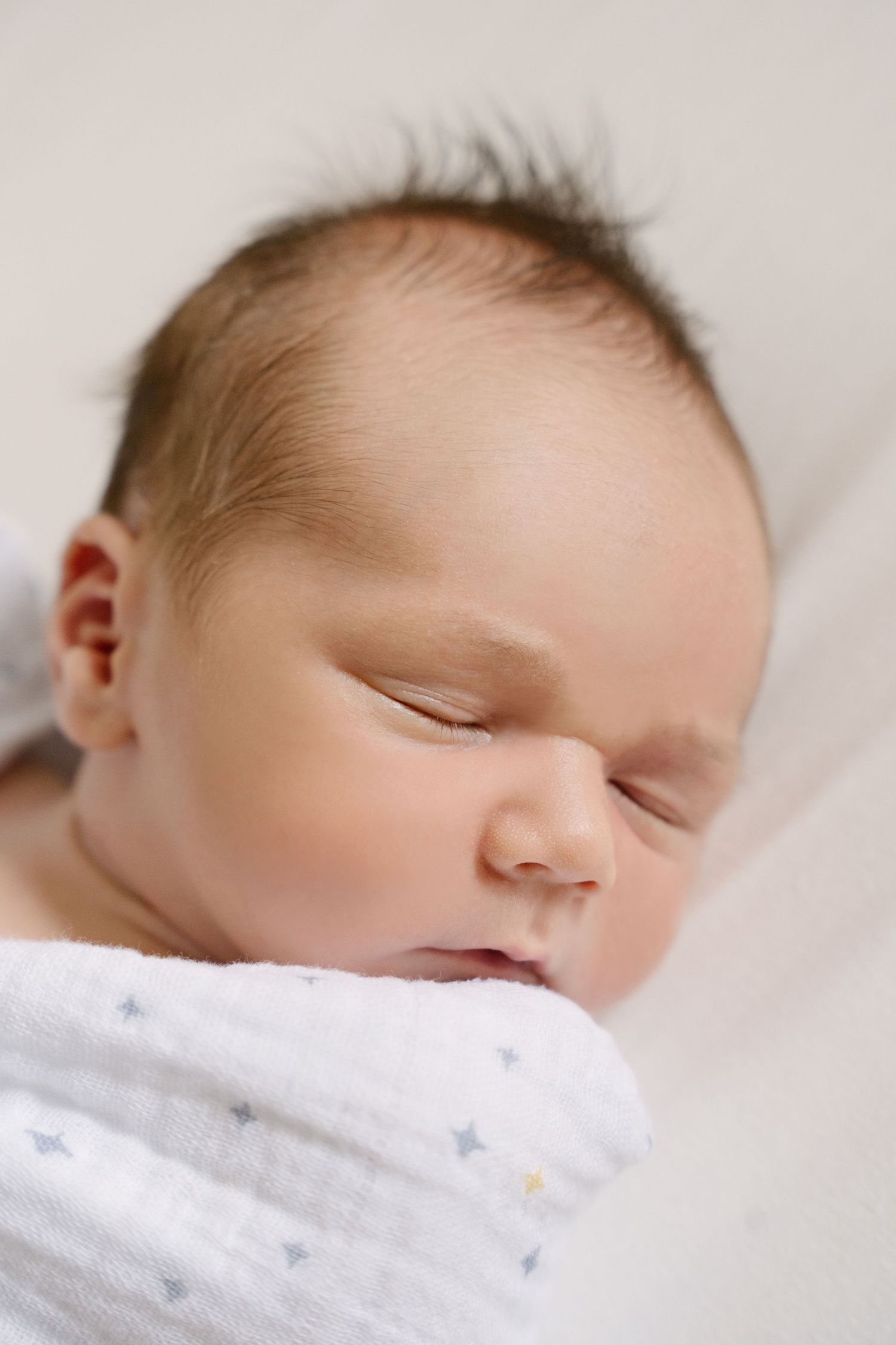 Peaceful newborn baby sleeps swaddled in a white star-print blanket in a soft, serene setting.