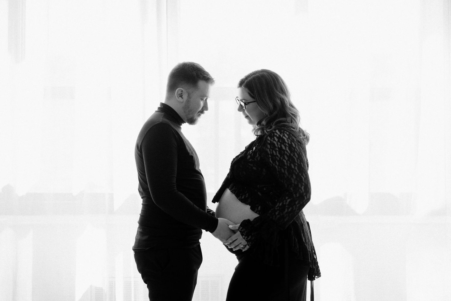 Black and white silhouette of couple holding hands facing each other by bright window.