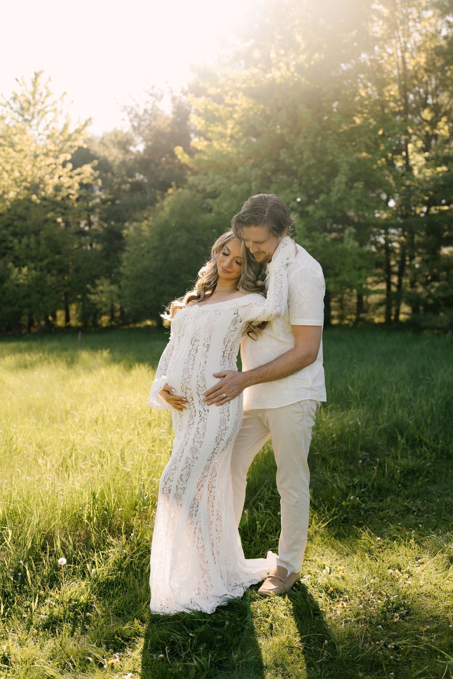 Man tenderly embracing pregnant woman in white lace dress from behind in a sunlit green field.