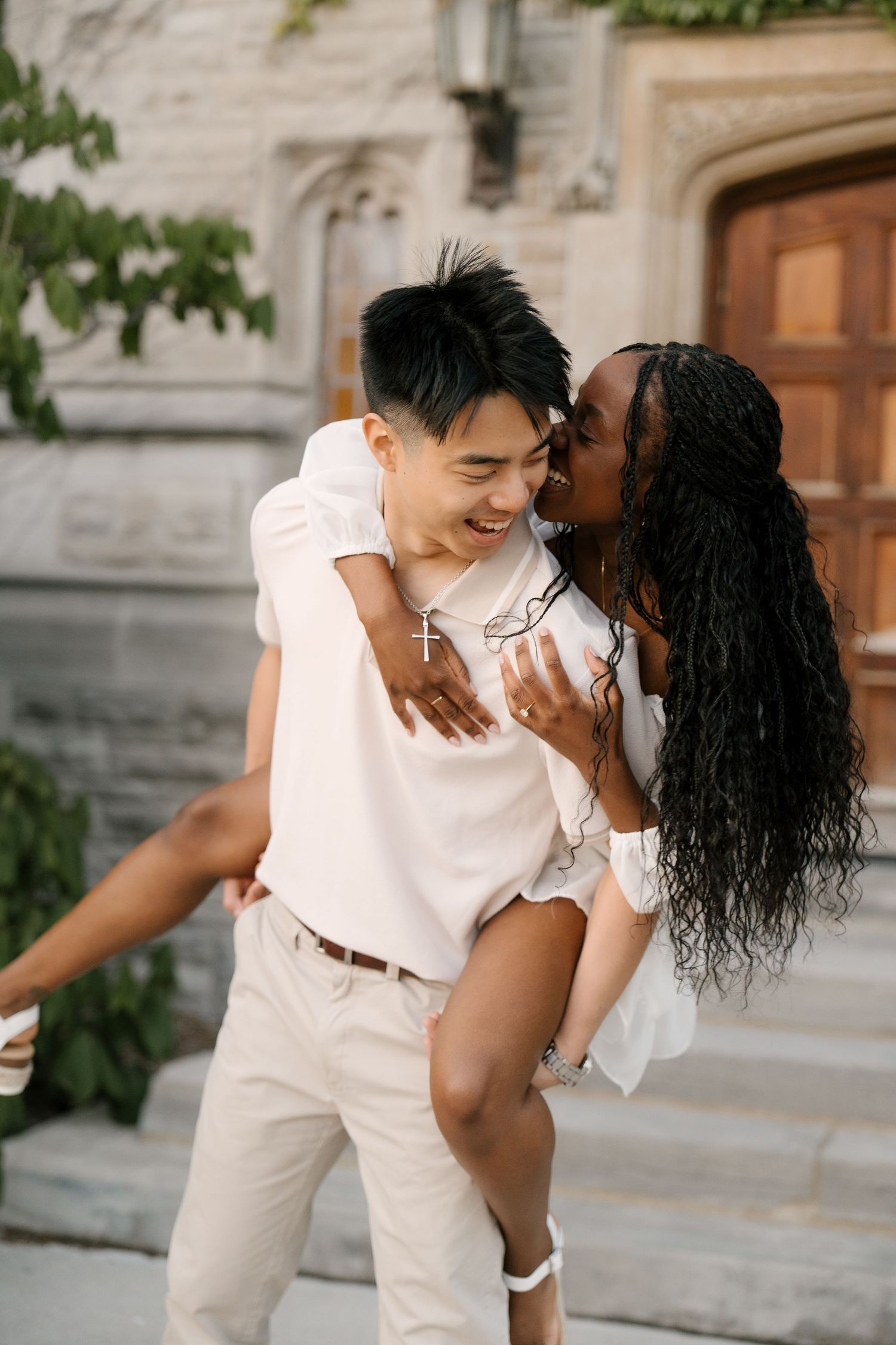 Man giving woman a piggyback ride while she kisses his cheek, laughing outside a stone building.