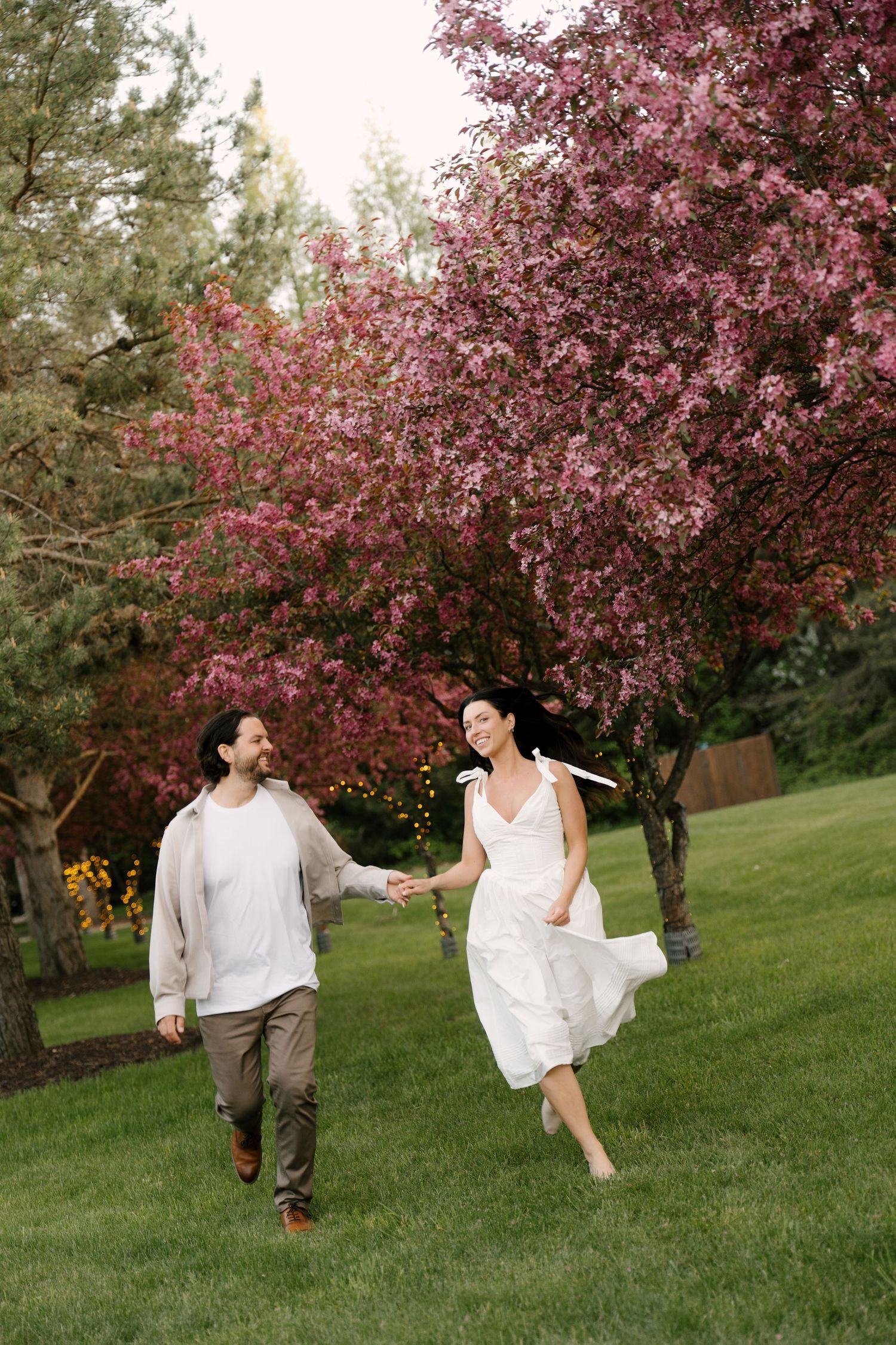 Joyful couple holding hands and running on green lawn beside a vibrant pink flowering tree.