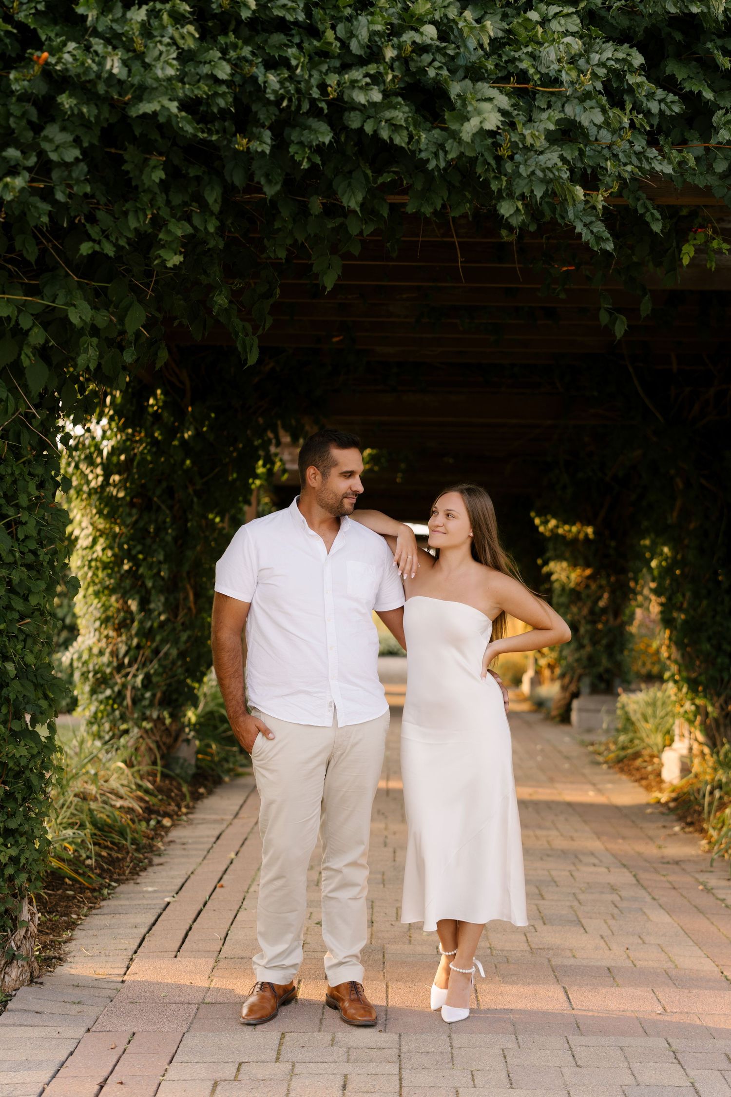 Couple walking together under green ivy pergola archway at sunset, woman in white dress leaning on man.