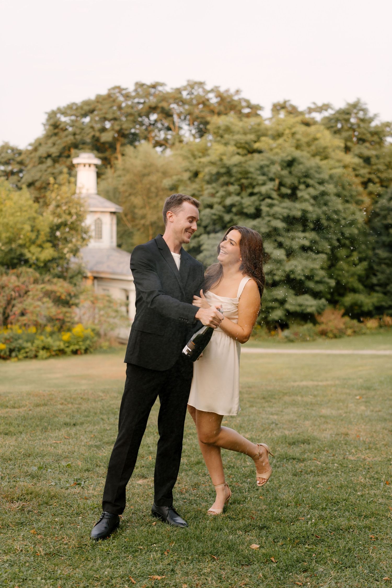 Happy couple dancing on green lawn popping champagne bottle with historic gazebo in background.
