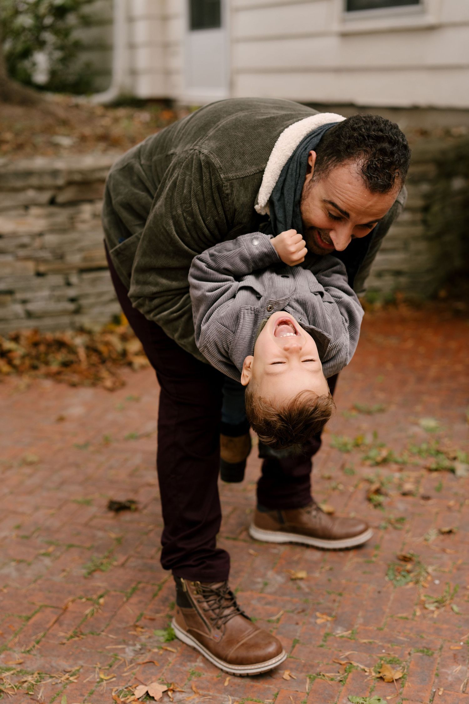 Playful father holding laughing toddler upside down on brick pathway in autumn setting.