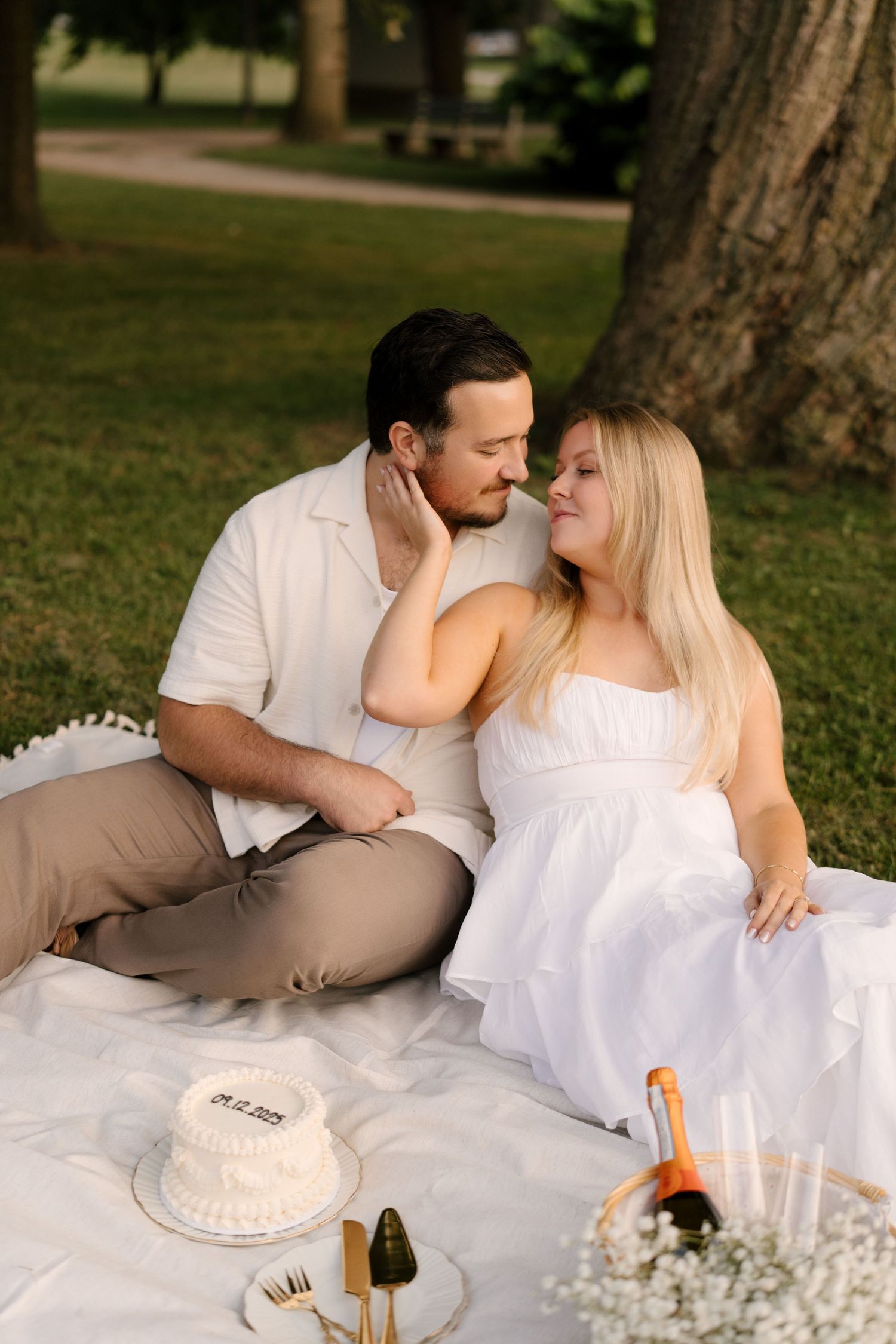 Couple sharing a romantic moment on a white blanket in a park with a wedding cake and champagne.