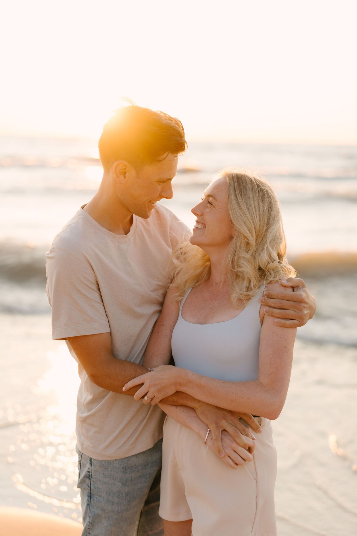 Happy couple embracing and laughing together on beach at warm golden sunset by the ocean.