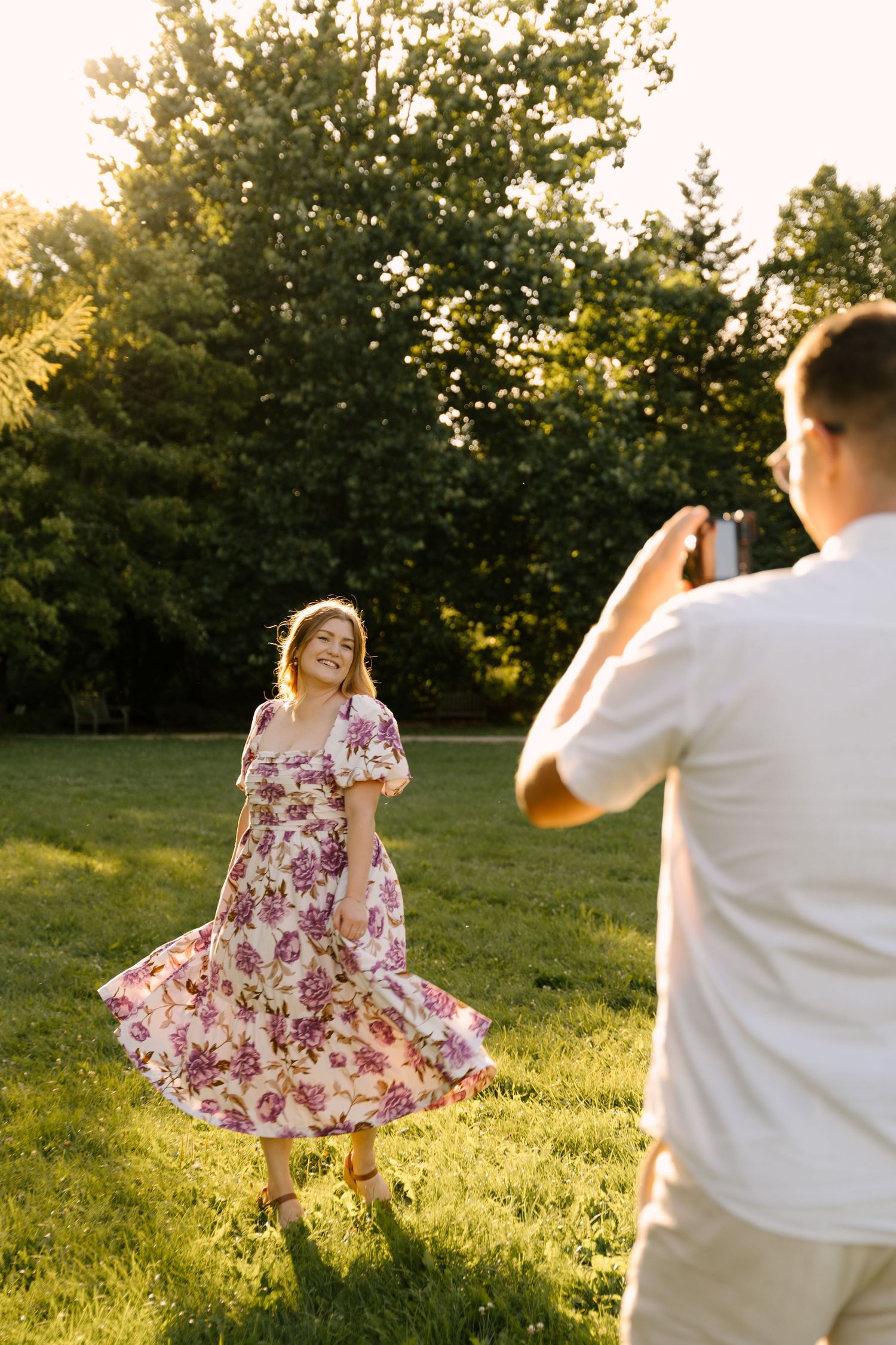 Man photographing smiling woman in floral dress twirling on green grass in golden sunlight.
