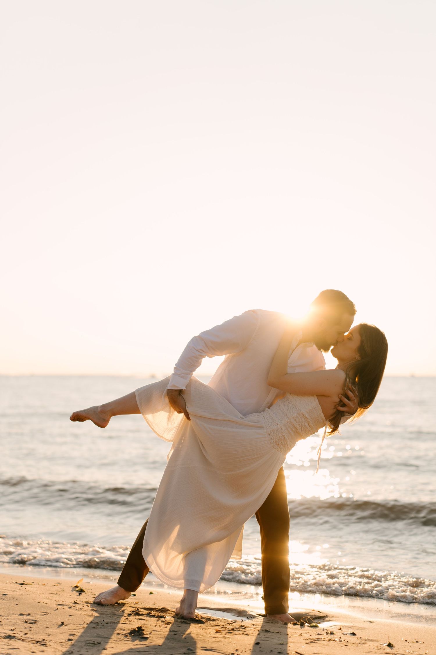 Couple sharing a passionate kiss on the beach at golden sunset, man dipping woman in white dress by the sea.