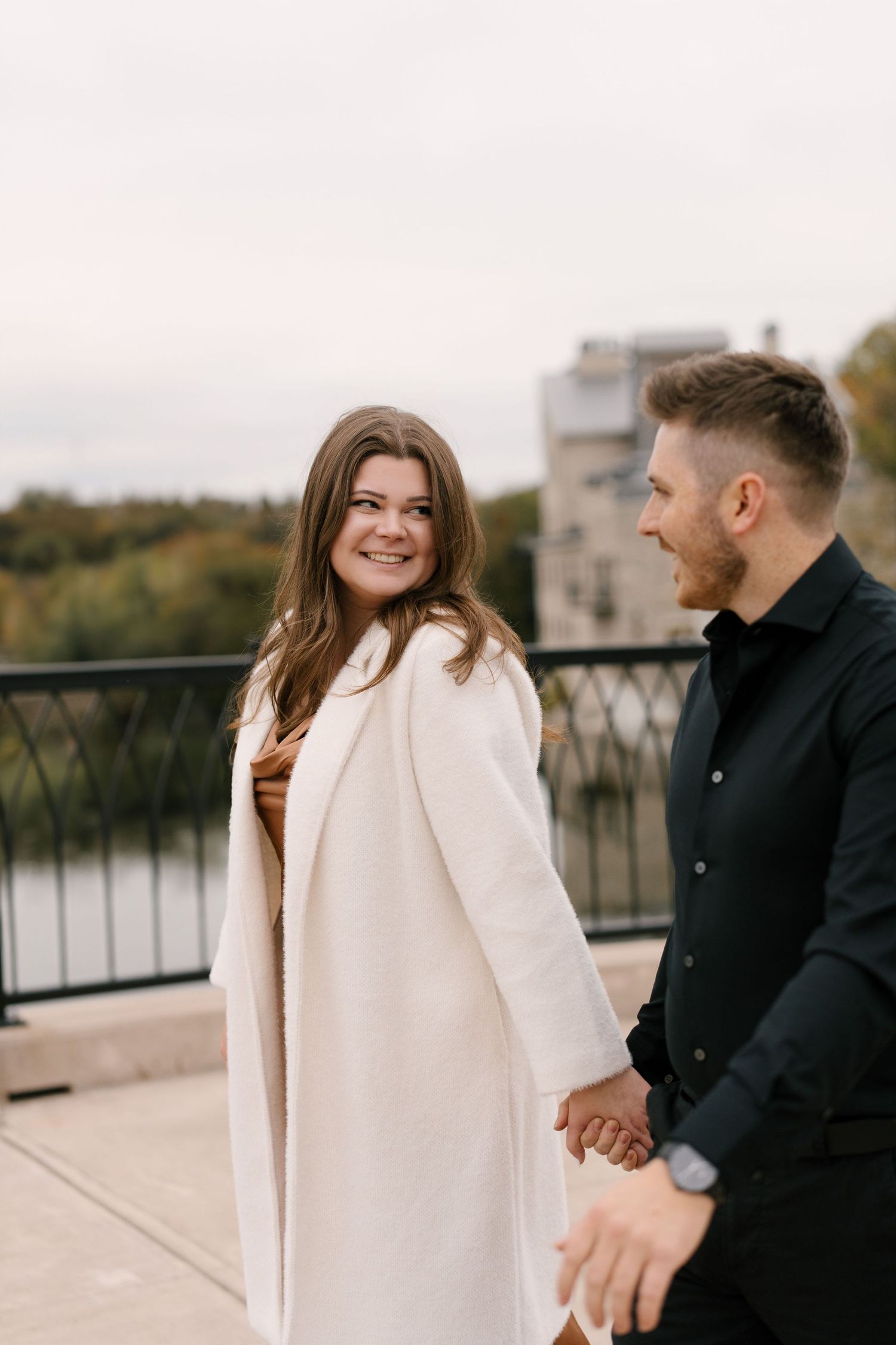 Smiling woman in white coat holds hands with man in black shirt during an autumn engagement photo session.