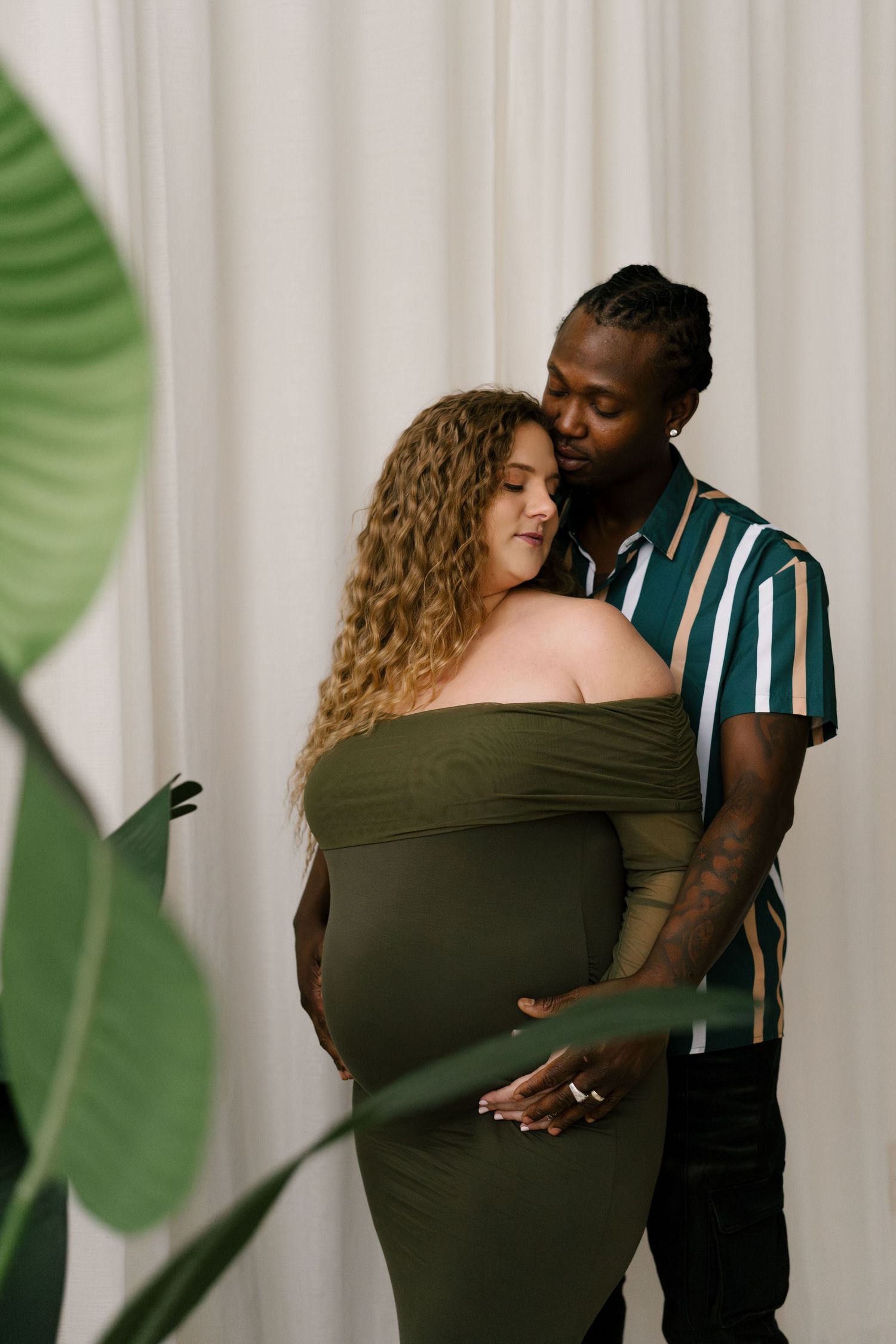 Interracial couple sharing tender moment embracing pregnant belly near tropical green leaves.