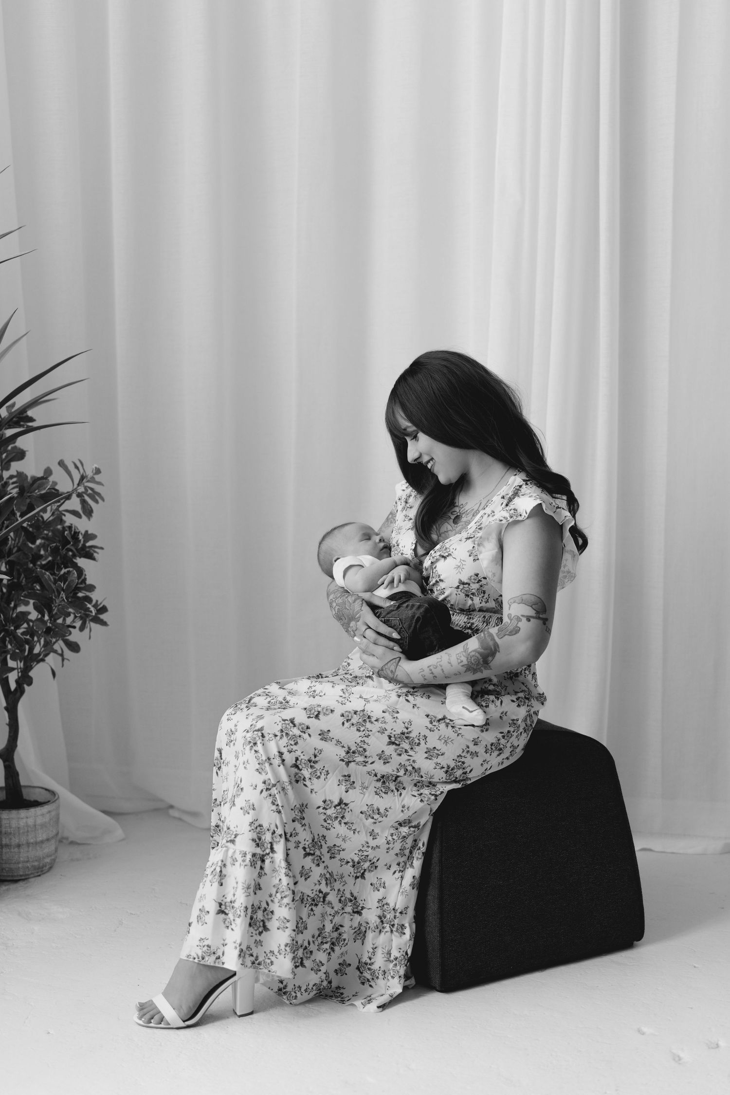 Black and white portrait of a tattooed mother in floral dress sitting and cradling her newborn baby.