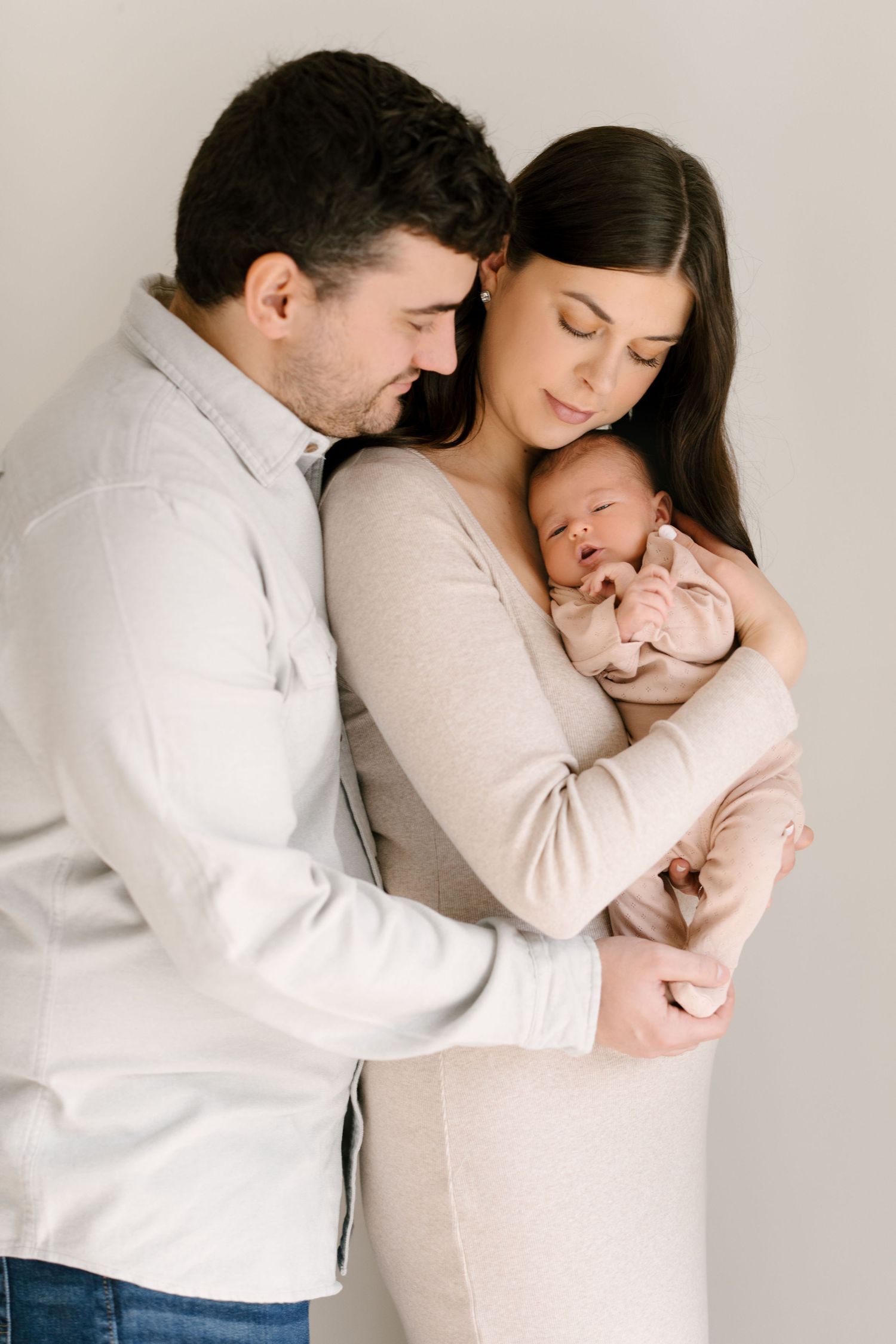 New parents tenderly cradling sleeping newborn baby dressed in pink against white background.