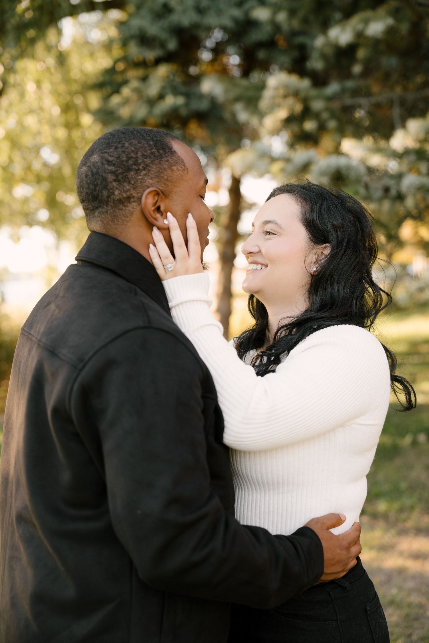 Smiling woman in white sweater tenderly holding man's face outdoors in soft natural light.