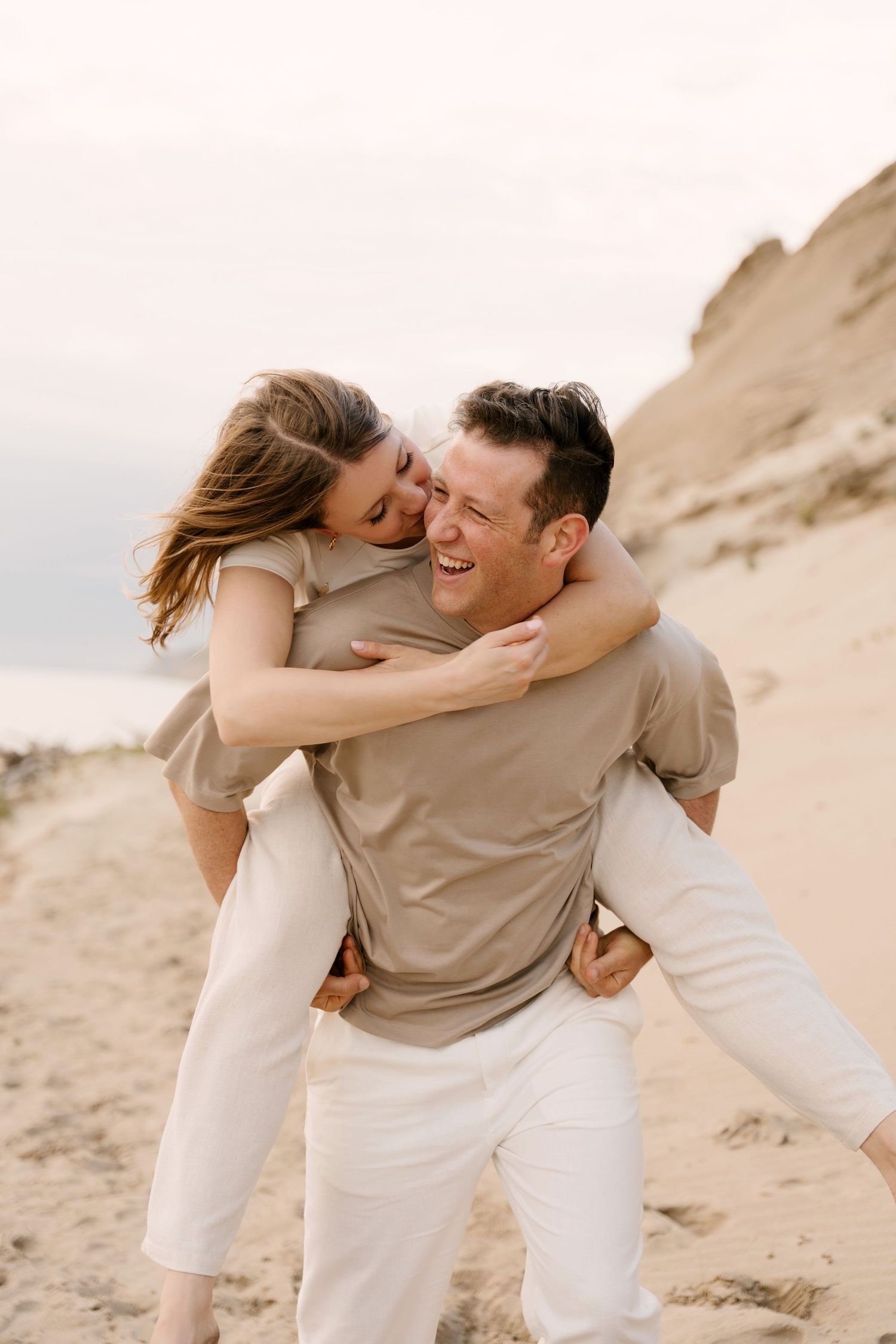 Man giving woman a piggyback ride on the beach, both laughing joyfully.