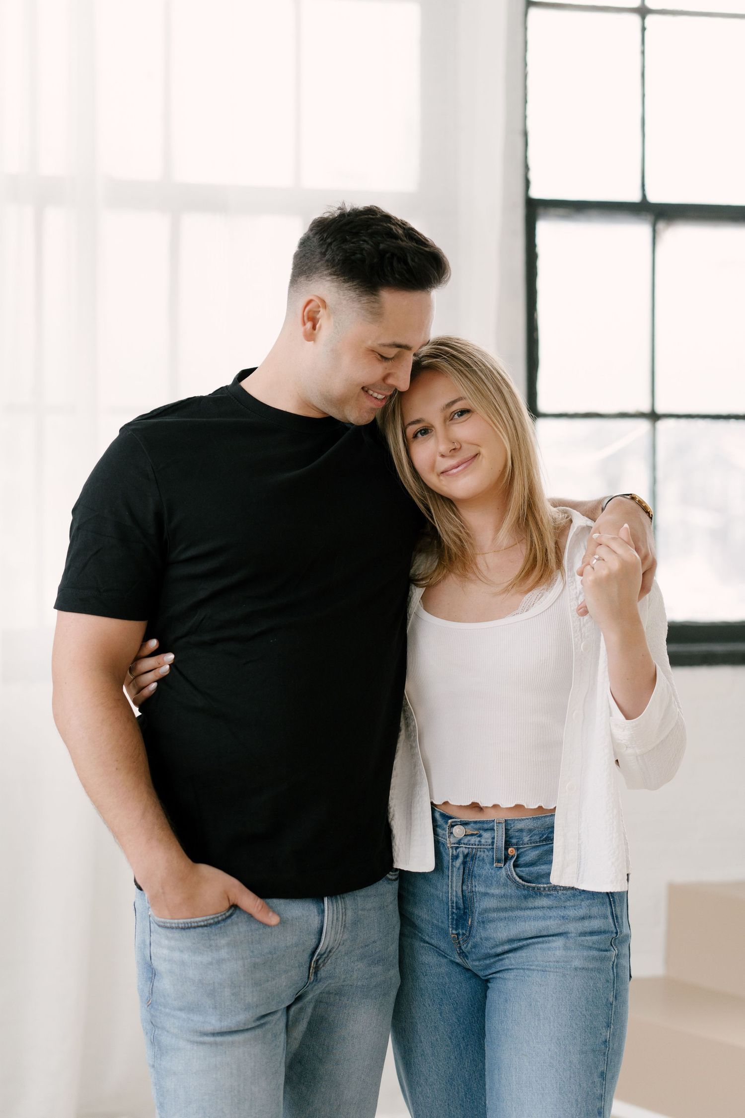 Happy blonde woman and dark-haired man in black tee holding hands tenderly in a bright room.