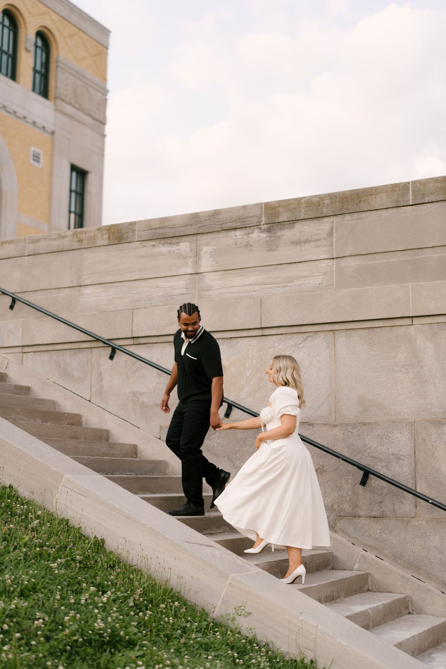 A couple walks hand in hand up outdoor stone steps beside a large concrete wall on a cloudy day.