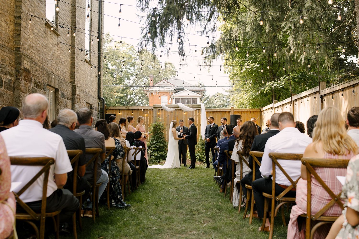 Outdoor wedding ceremony with guests seated on wooden chairs as couple exchanges vows under string lights.