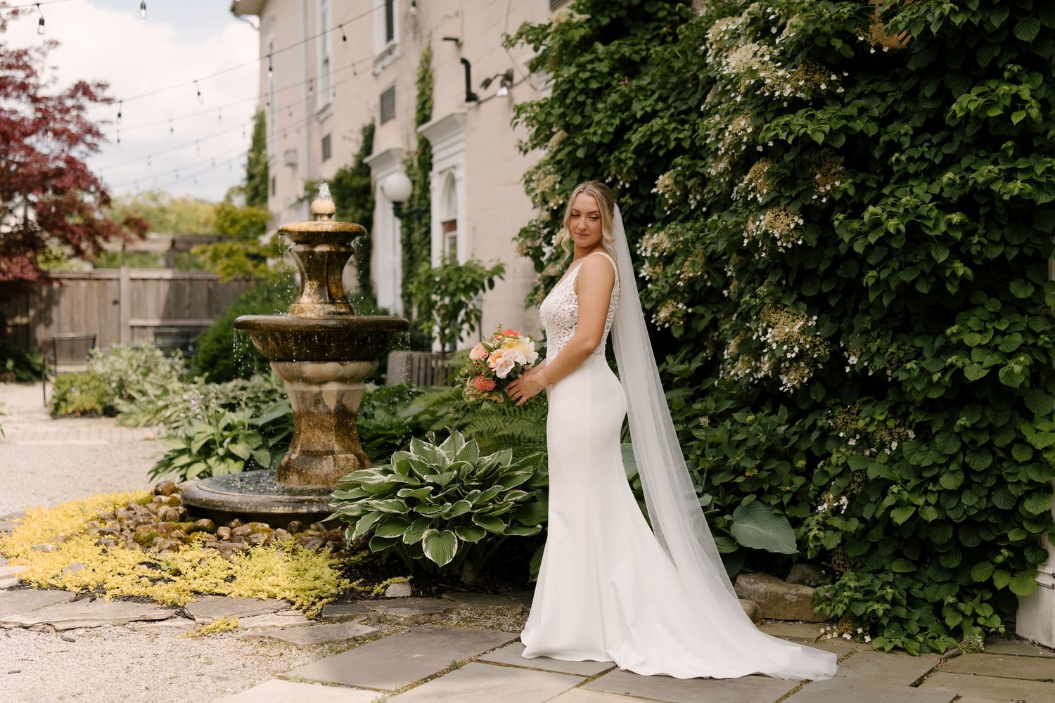 Elegant bride in fitted gown poses beside stone fountain in lush garden setting at Inn On The Twenty.