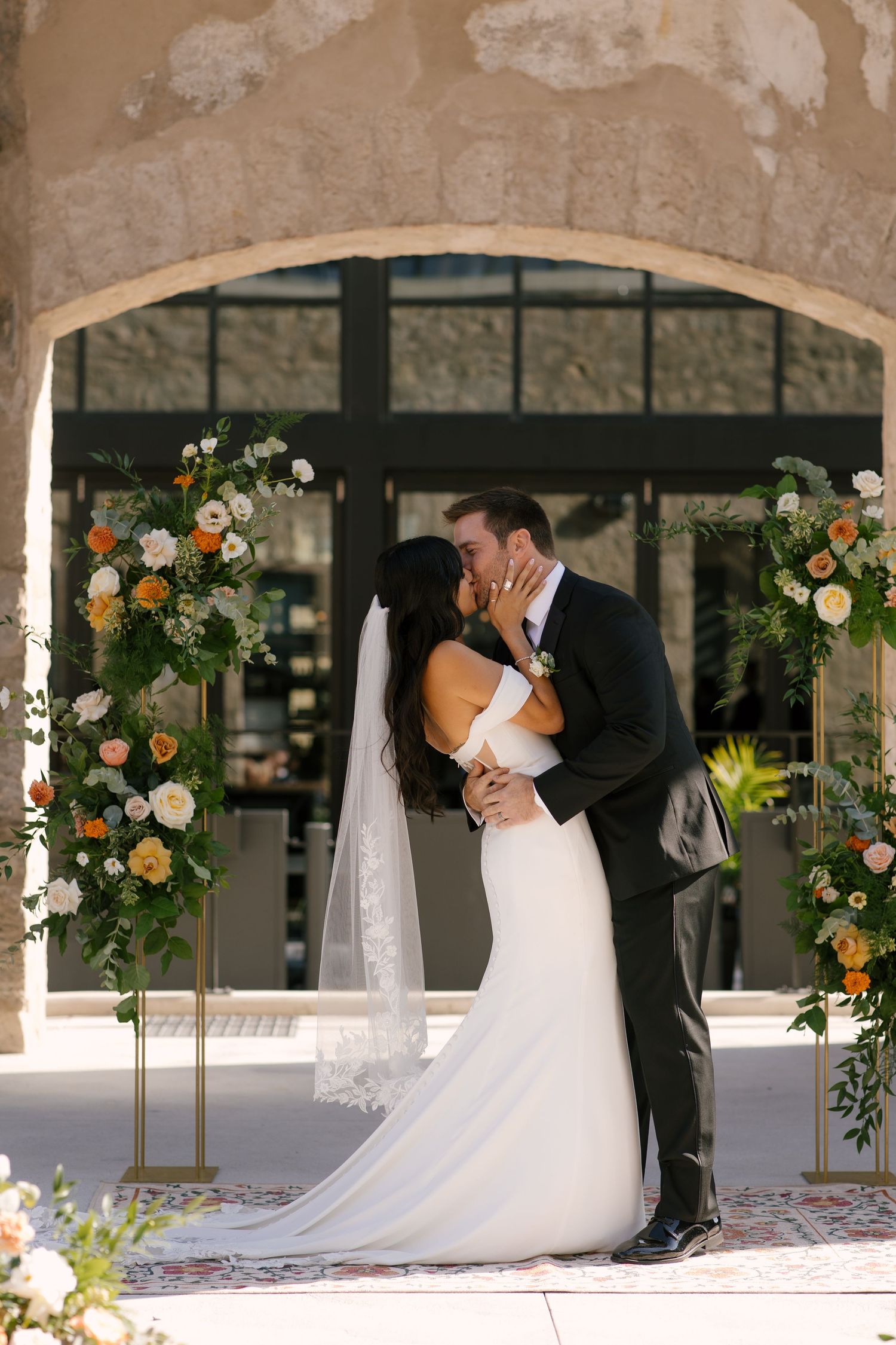 Bride and groom share first kiss at outdoor Tapestry Hall ceremony altar adorned with orange and white floral arrangements.