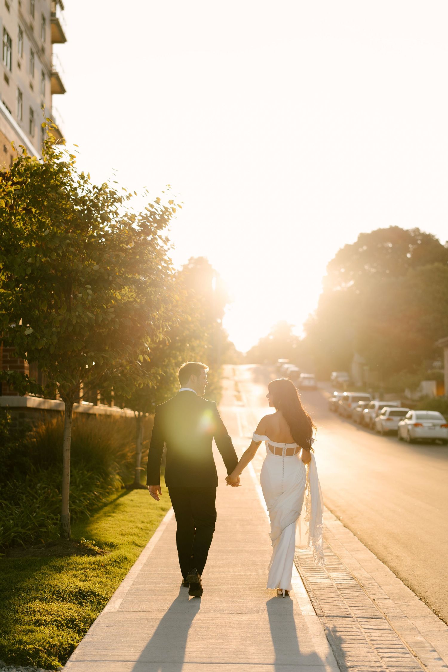Bride and groom walking hand in hand into radiant golden sunset on a tree-lined urban sidewalk outside Tapestry Hall.