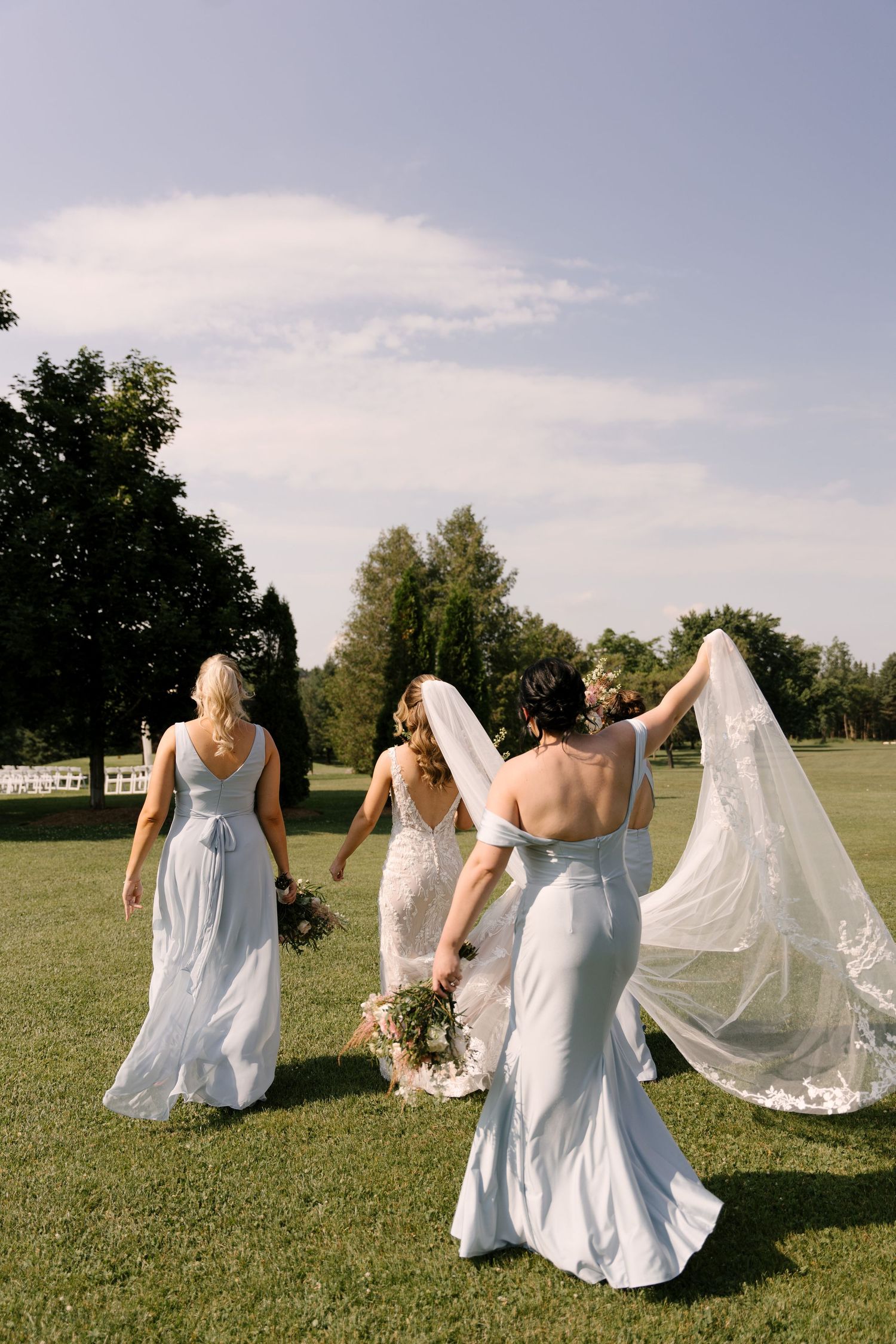 Bride and bridesmaids in light blue gowns walking across a green lawn on a sunny wedding day at St. Marys Wedding Venue.