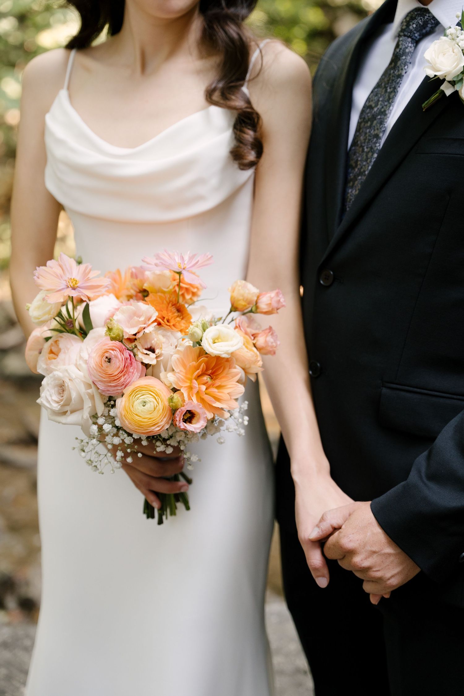 Bride and groom holding hands, bride wearing white gown holding peach and pink floral bouquet, groom in black suit.