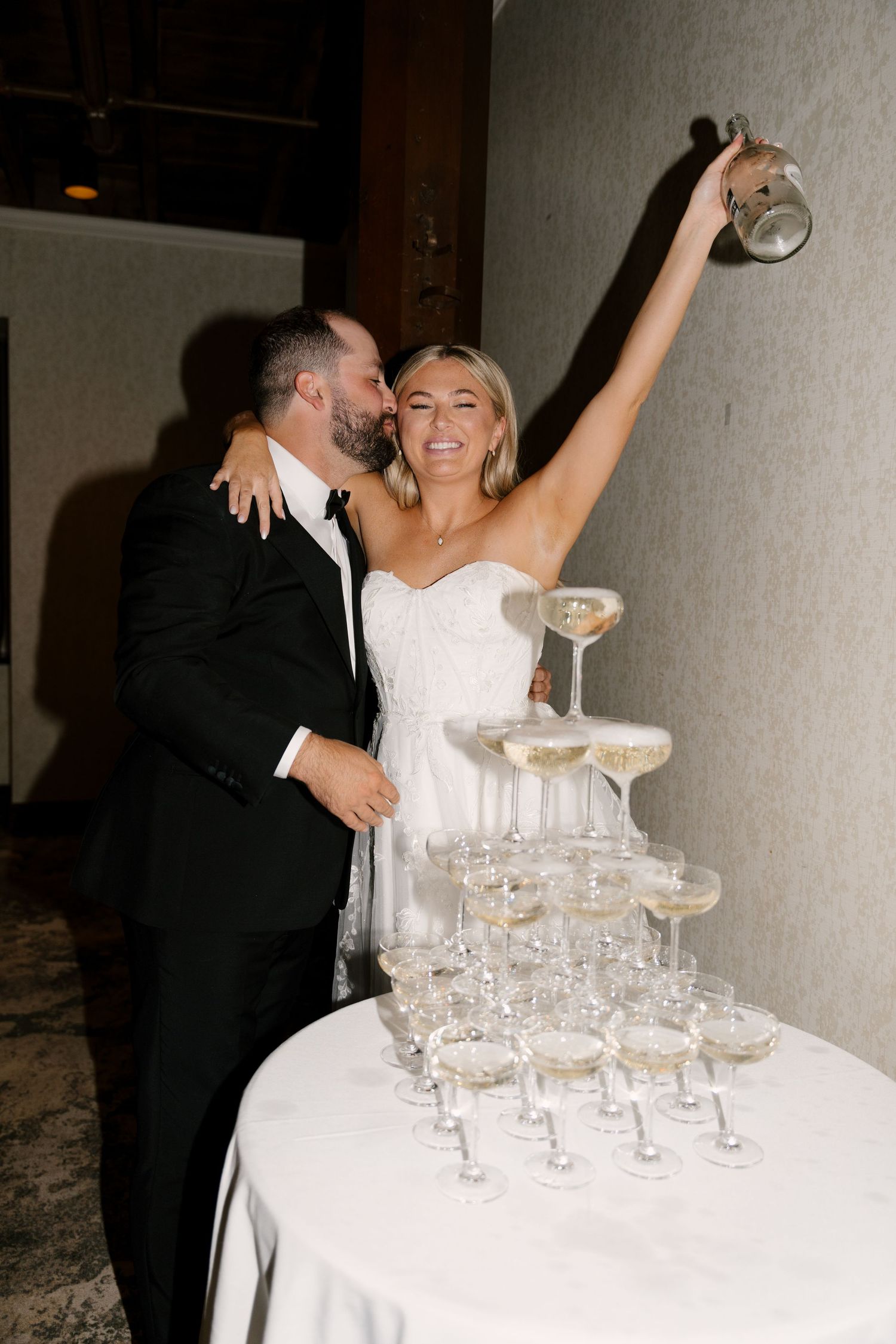 Bride raises champagne bottle over elegant tower of coupe glasses while groom embraces her at wedding reception.