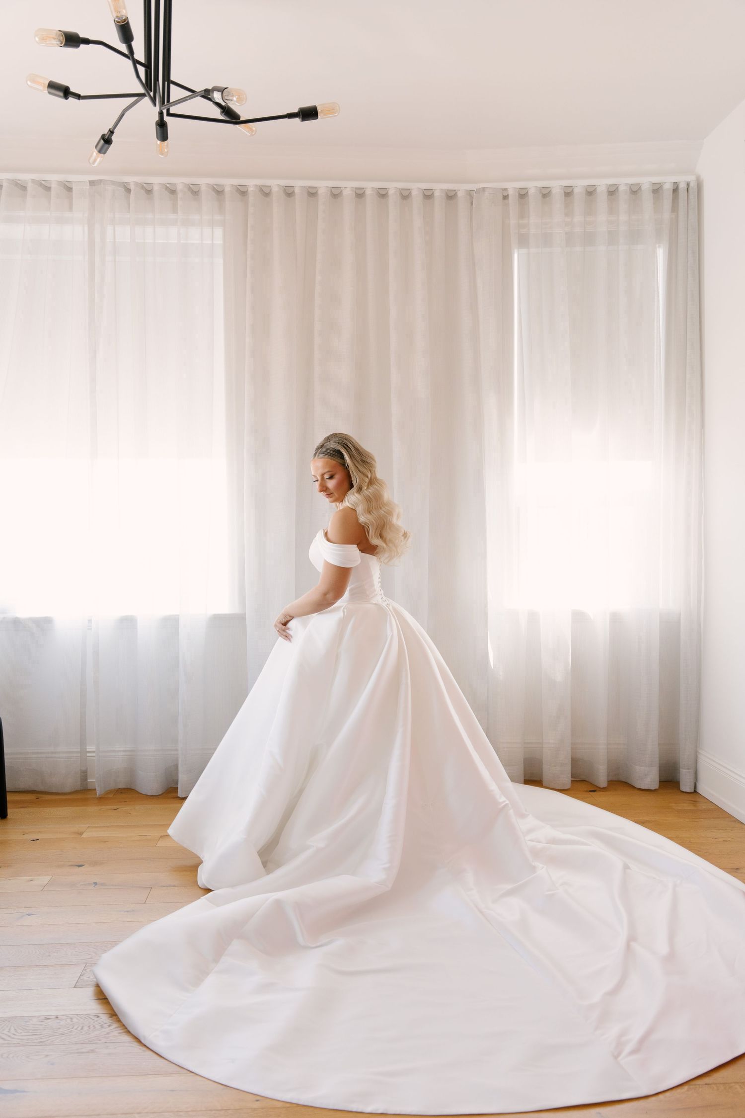 Bride in off-shoulder ballgown with long train standing by sheer curtained windows in The Walper Hotel.