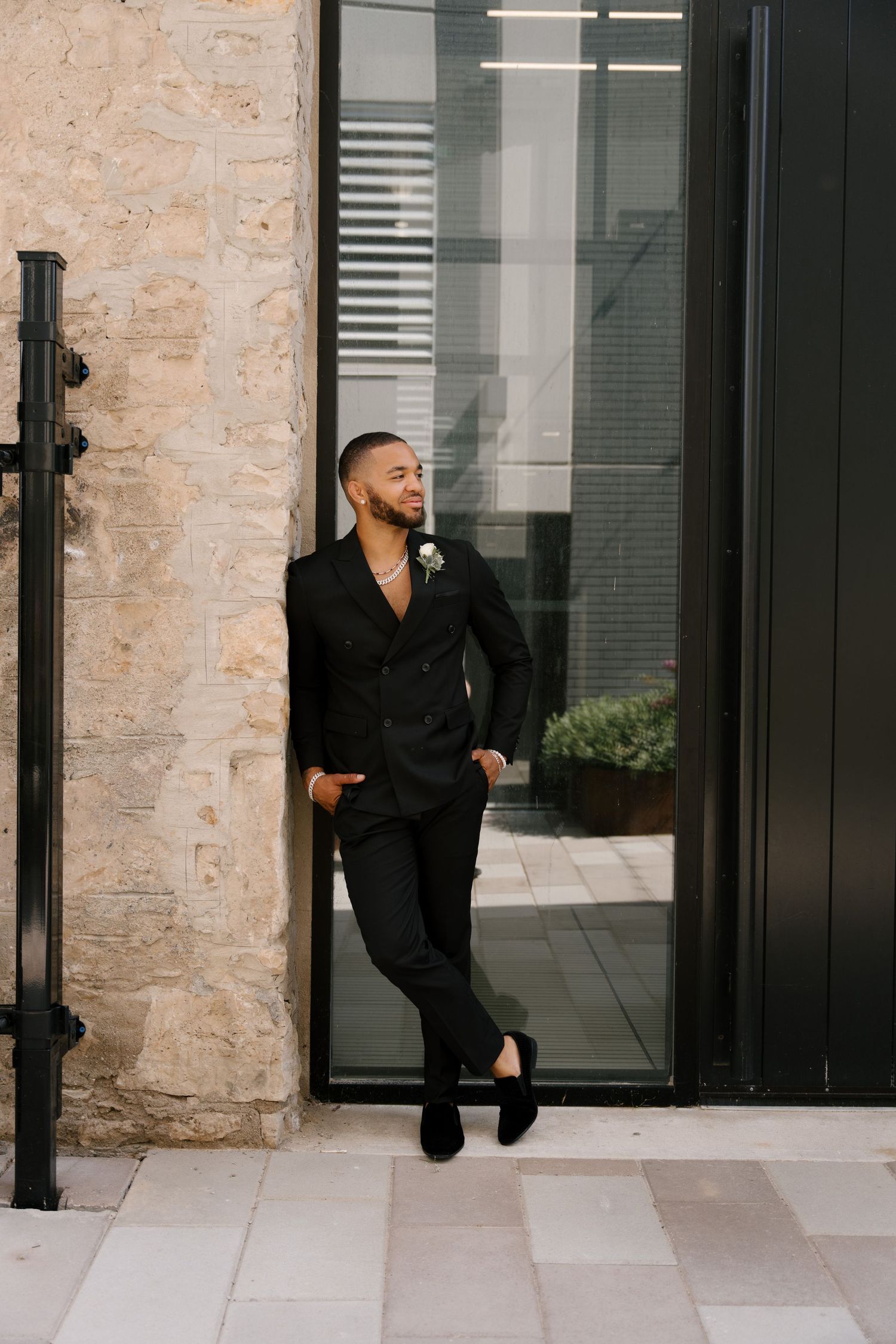 Stylish groom in a black double-breasted suit leans against a stone wall outside a modern Tapestry Hall wedding venue.