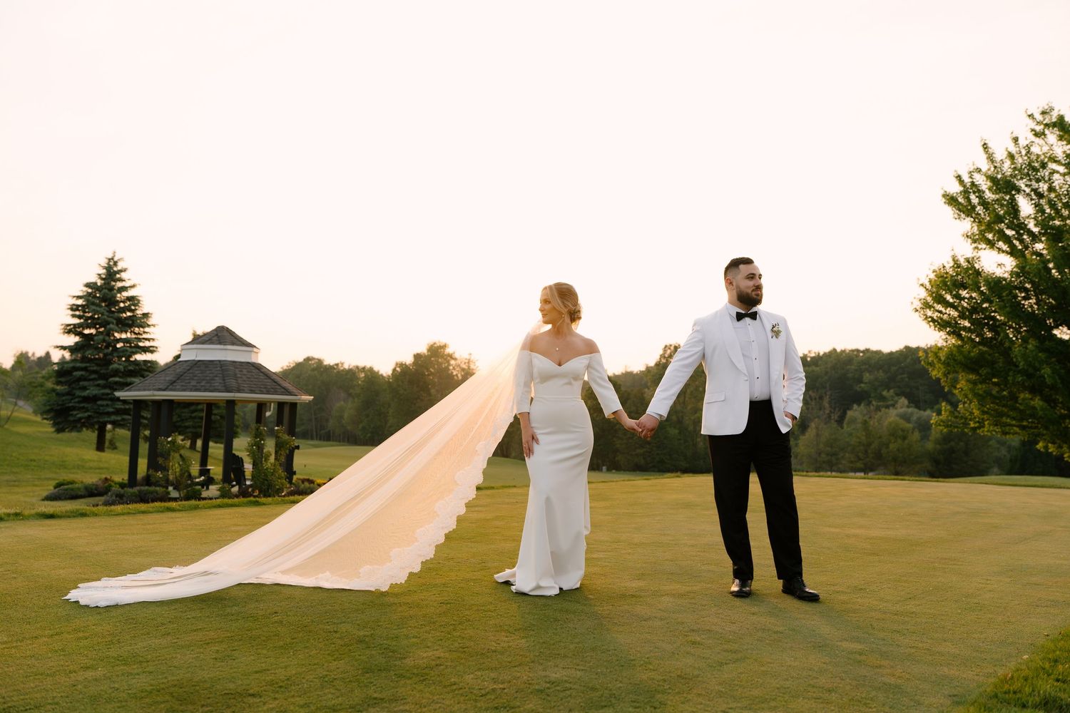 Bride and groom holding hands on lush Whistle Bear golf course at dusk, long cathedral veil trailing elegantly behind bride.