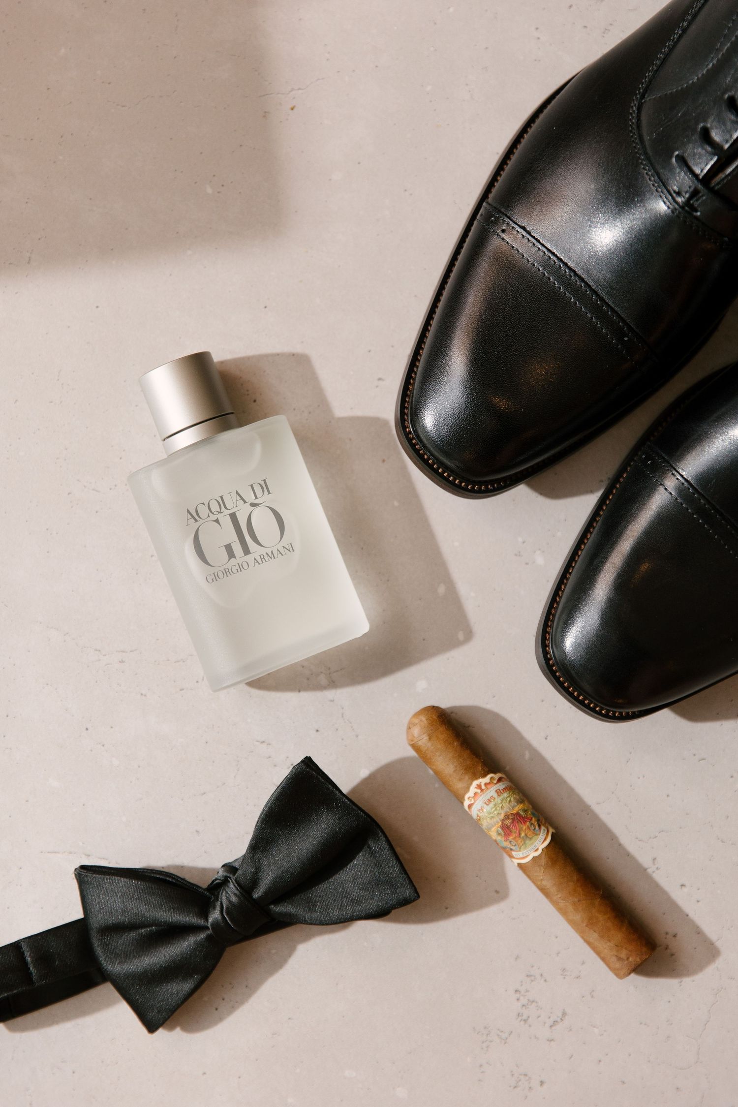 Wedding day groom details flat lay with Acqua di Gio cologne, black bow tie, cigar, and dress shoes.