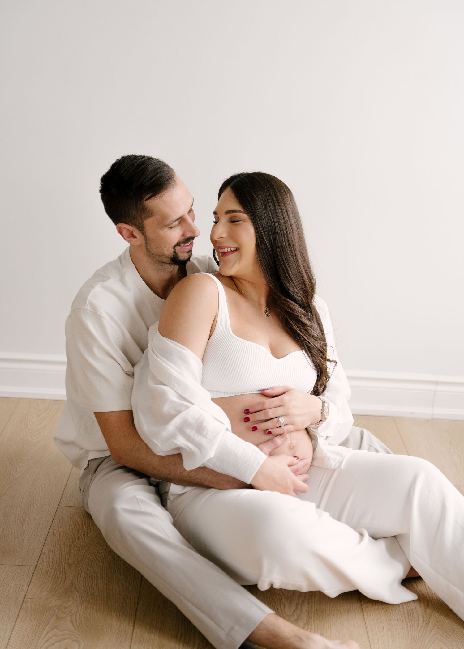 Happy couple in matching white outfits sitting on floor, man hugging smiling pregnant woman in bright studio.