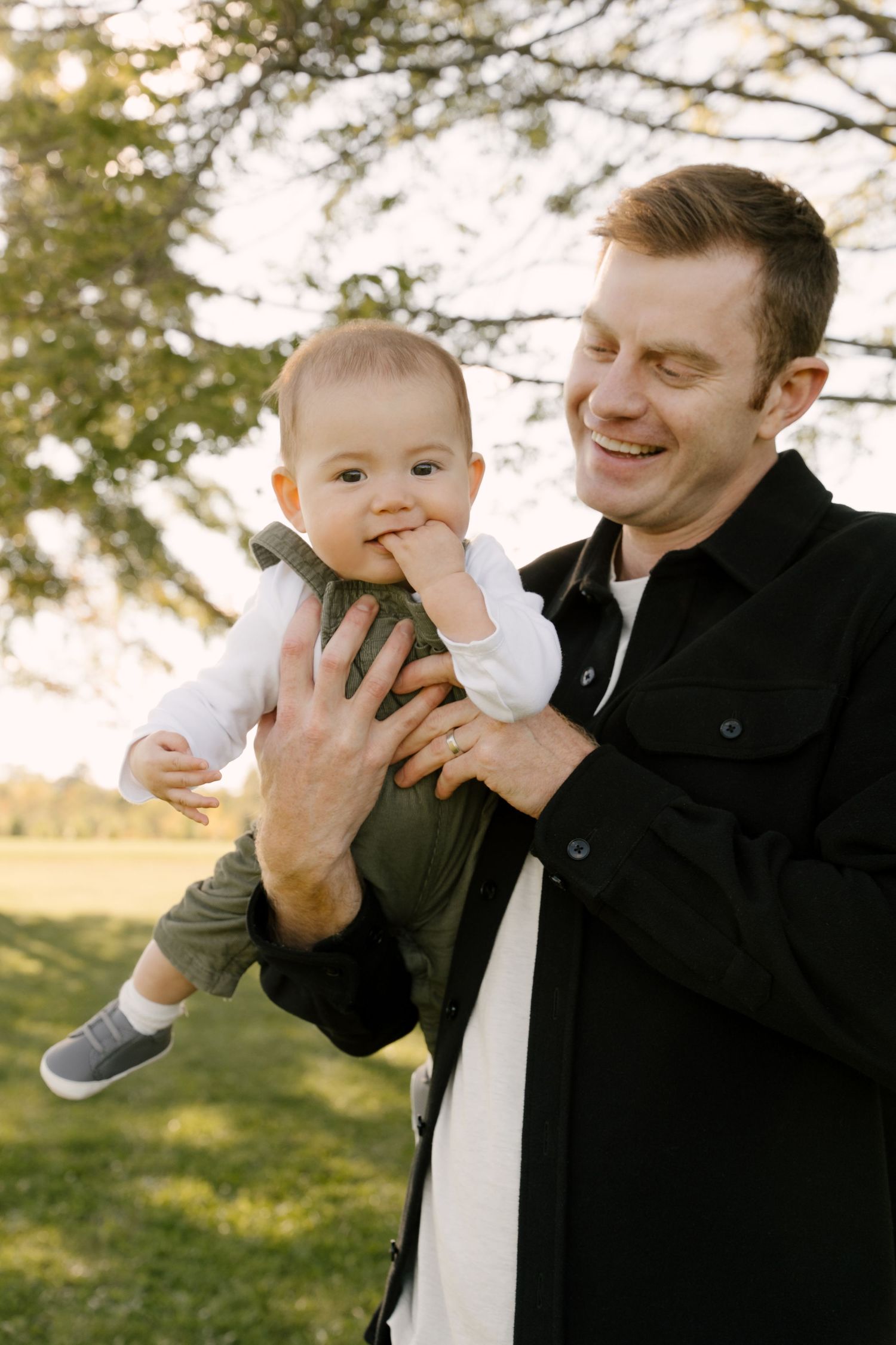 Smiling father holding baby in olive overalls outdoors in sunny park setting.
