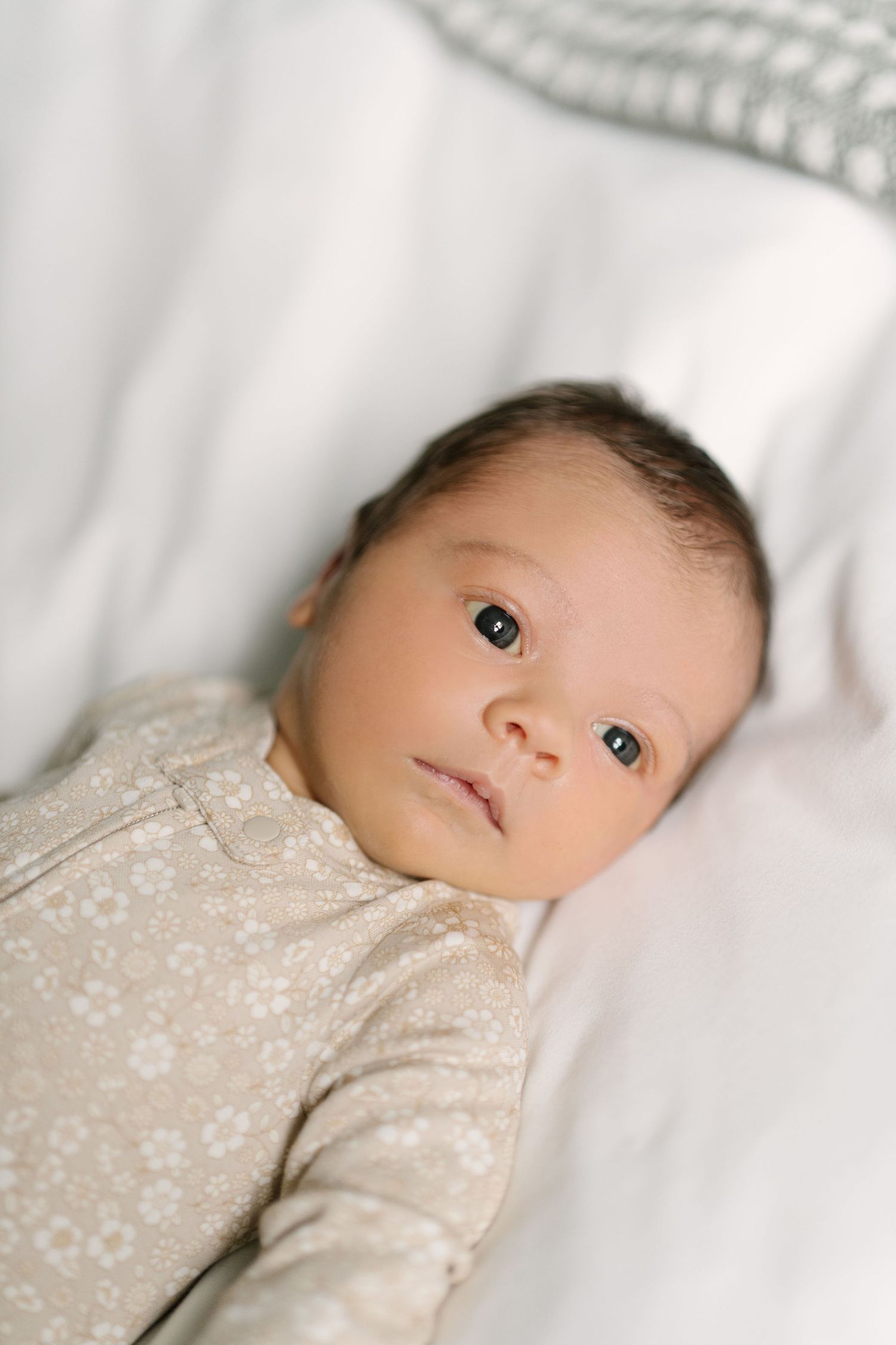 Newborn baby with dark eyes lying on white bedding wearing a beige floral onesie, looking up peacefully.