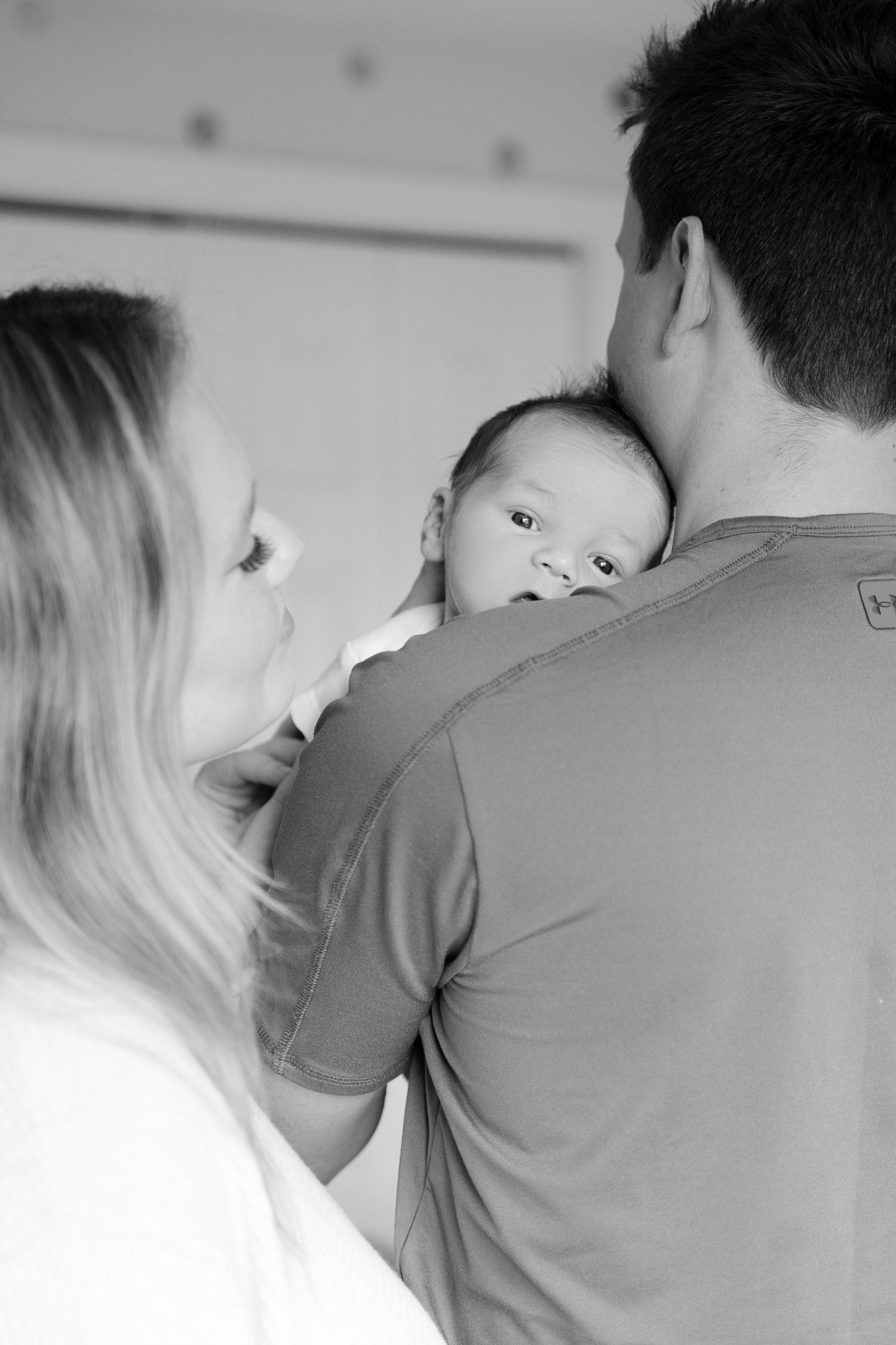 Black and white photo of alert newborn baby looking over fathers shoulder while mother watches.