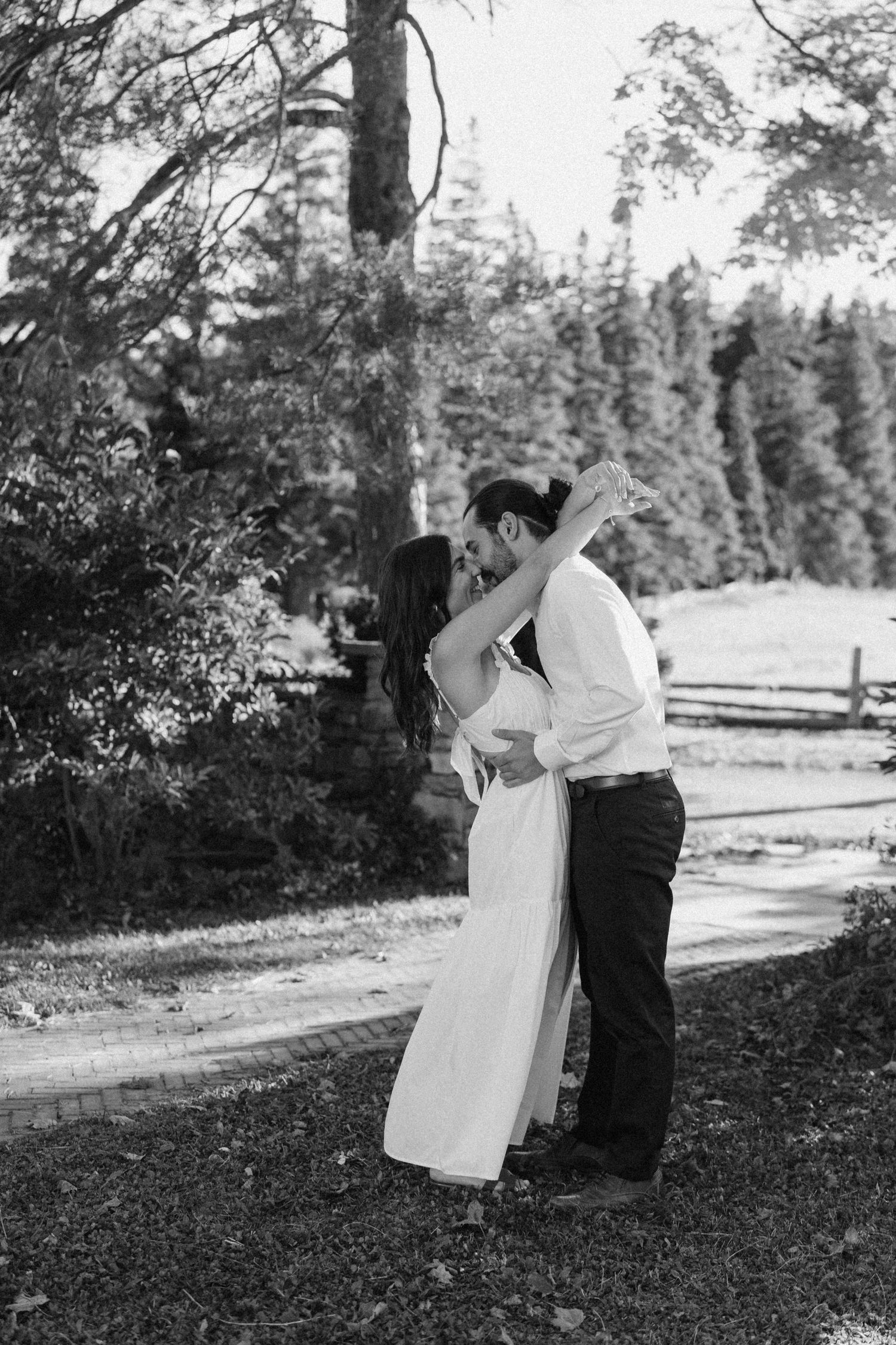 Black and white photo of couple sharing a romantic kiss on a tree-lined path.
