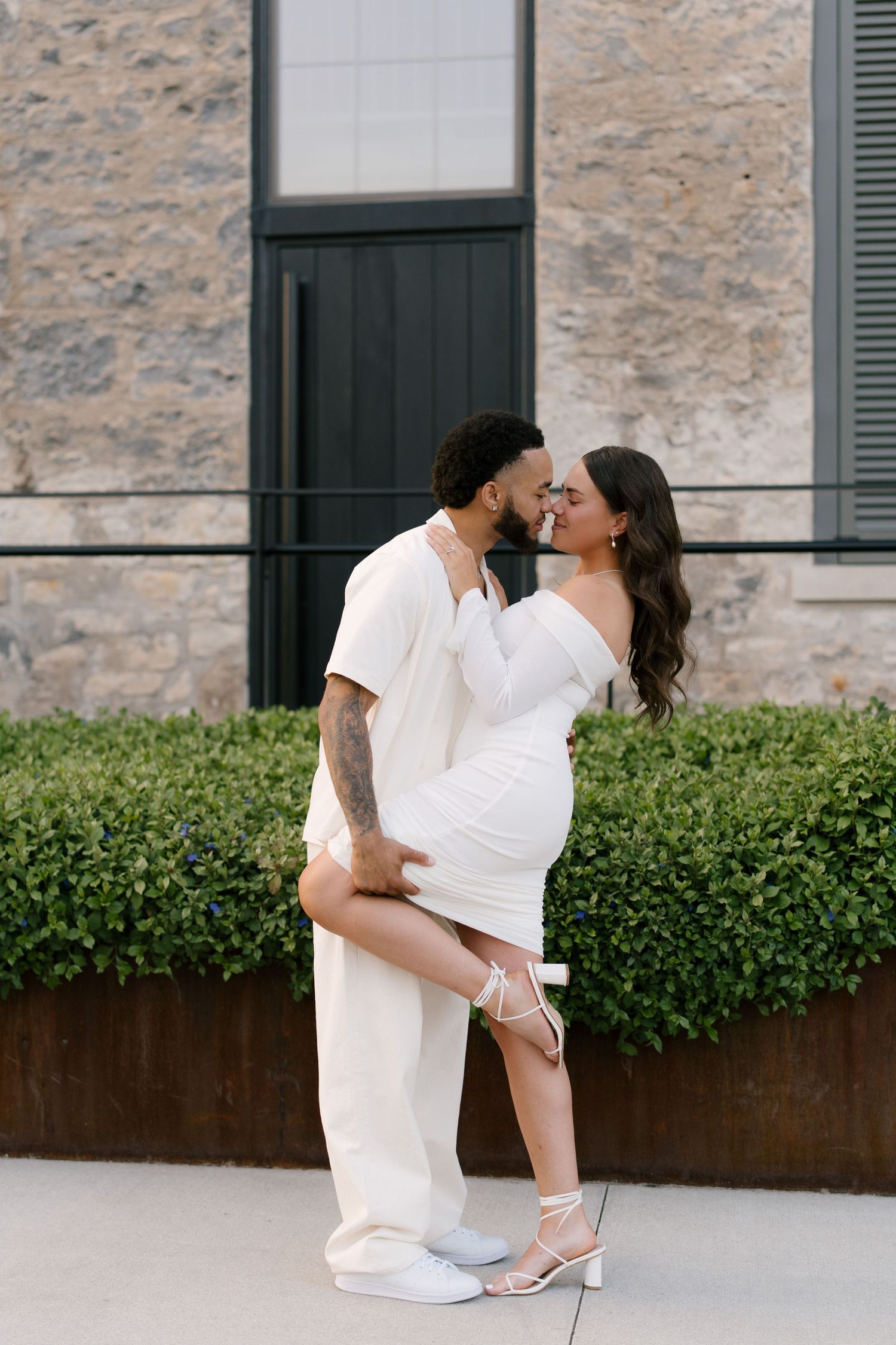 Couple in white outfits share a kiss outdoors, man lifts woman's leg romantically.