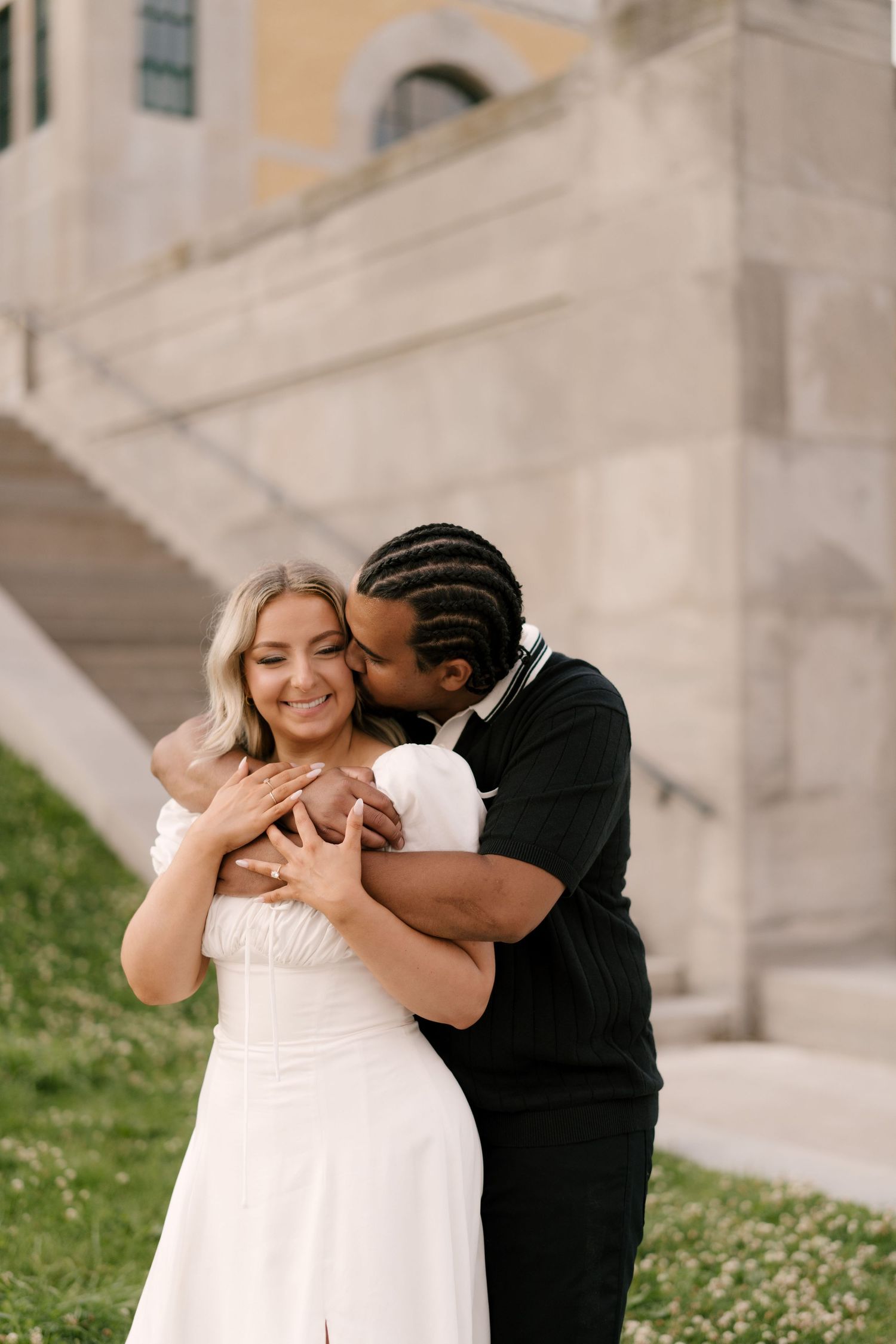 A man in black kisses a smiling woman in a white dress while embracing her near stone steps outdoors.
