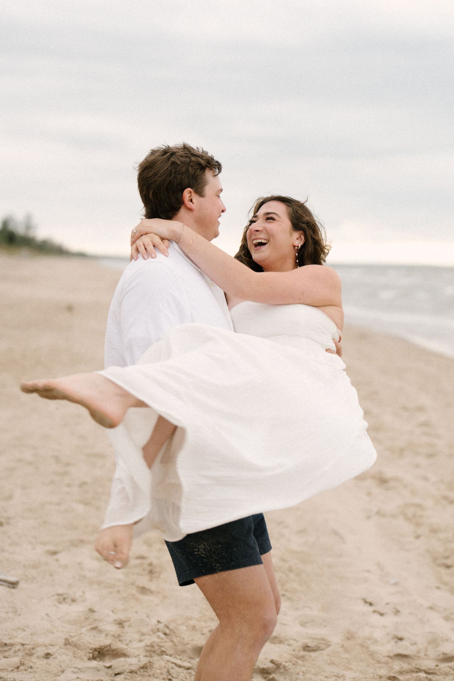 Groom lifting laughing bride in white dress on a sandy beach with ocean in the background.
