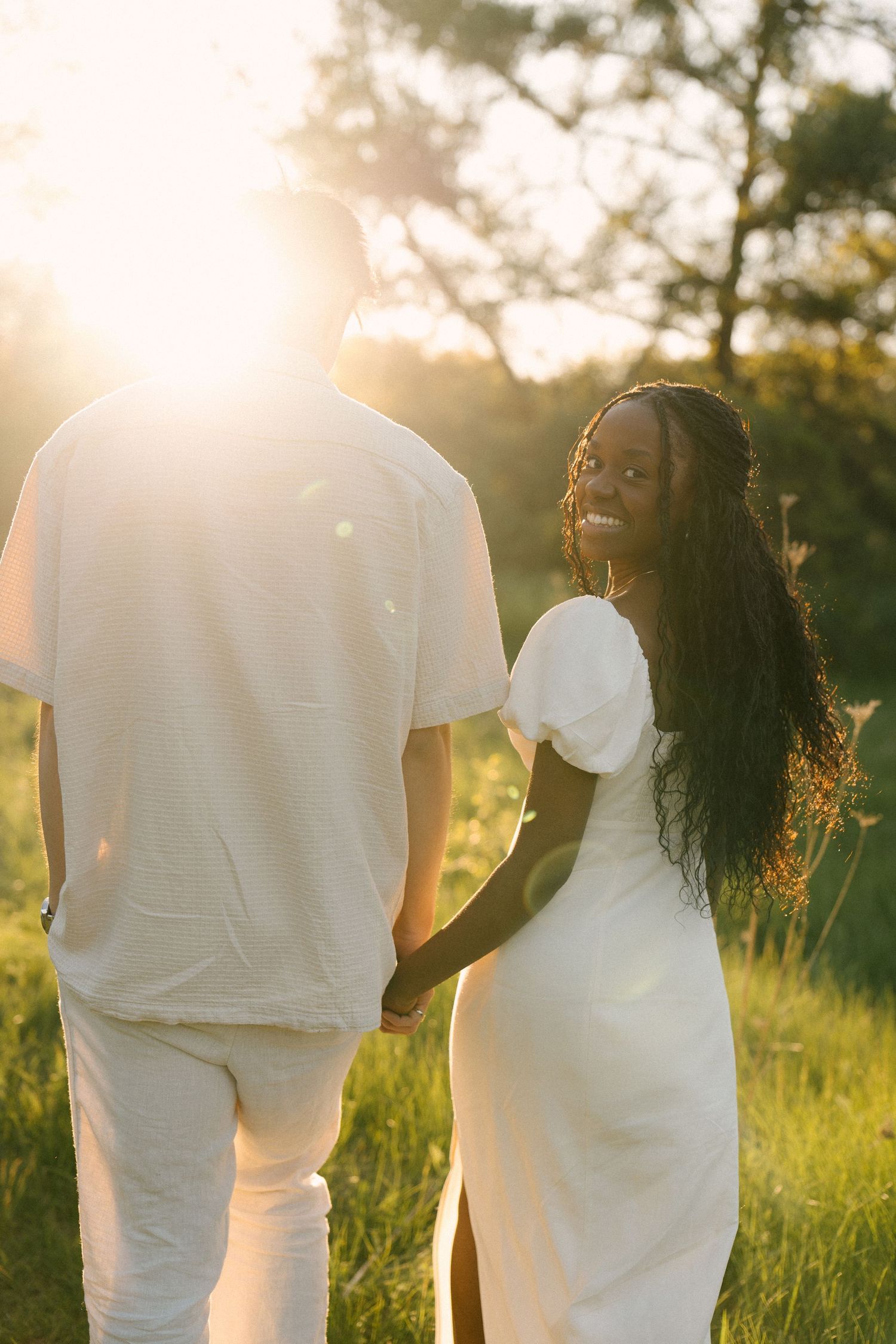 Smiling woman in white dress holds hands with partner walking in sunset field.