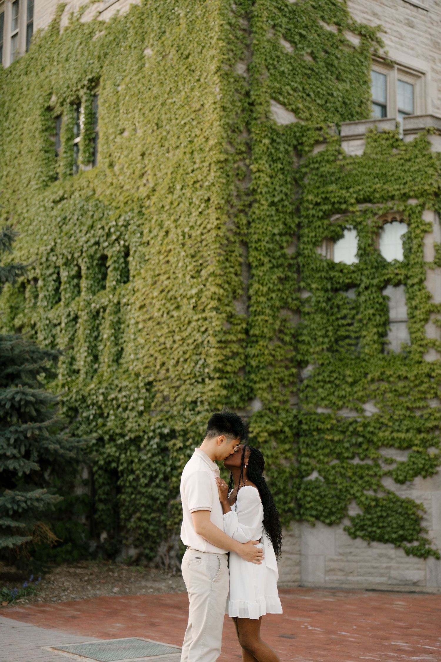 Interracial couple embracing and kissing in front of an ivy-covered stone building on a brick pathway.