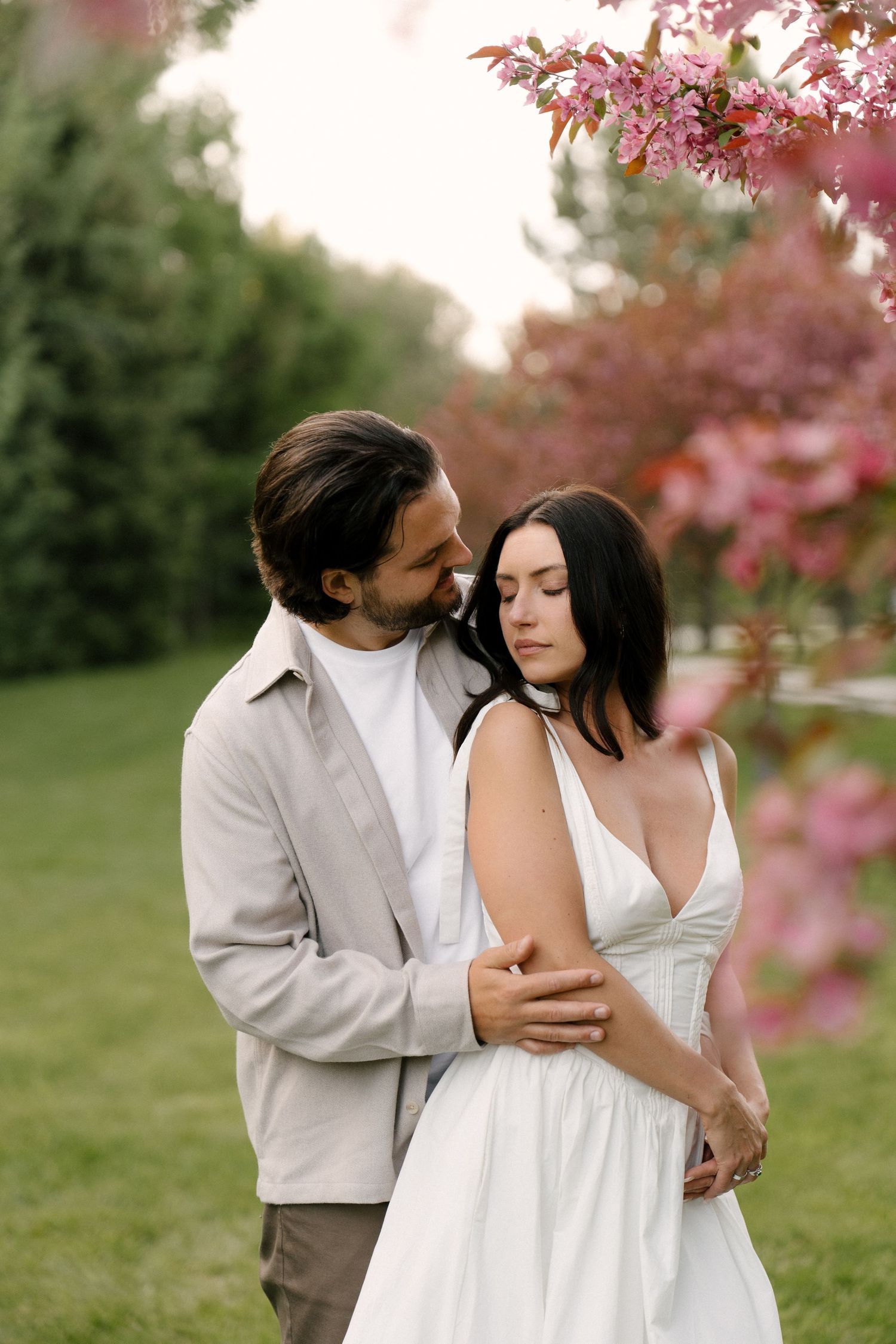 Couple sharing intimate embrace under pink flowering tree branches in soft natural light.