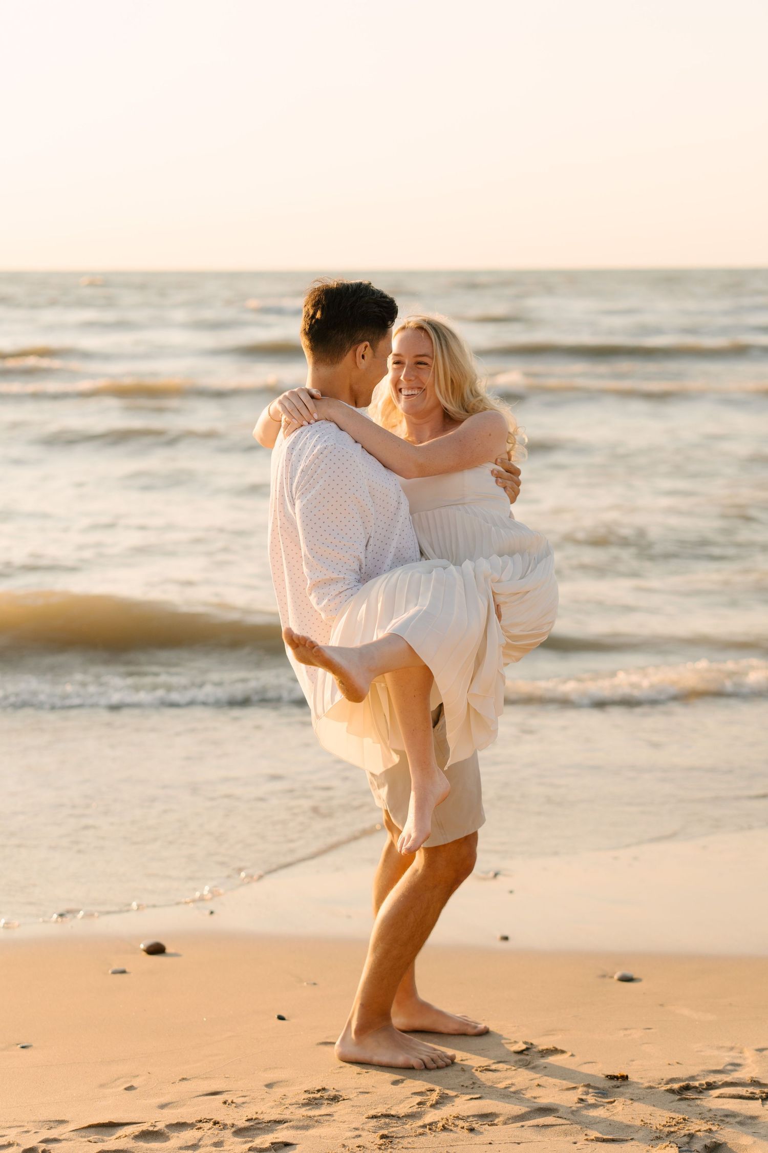 Man lifting smiling blonde woman in white dress on sandy beach at warm golden sunset.