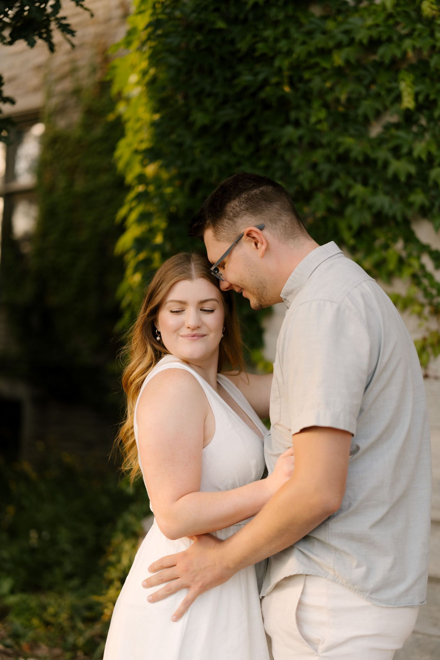 Couple embracing tenderly in front of lush ivy-covered stone wall in soft golden light.