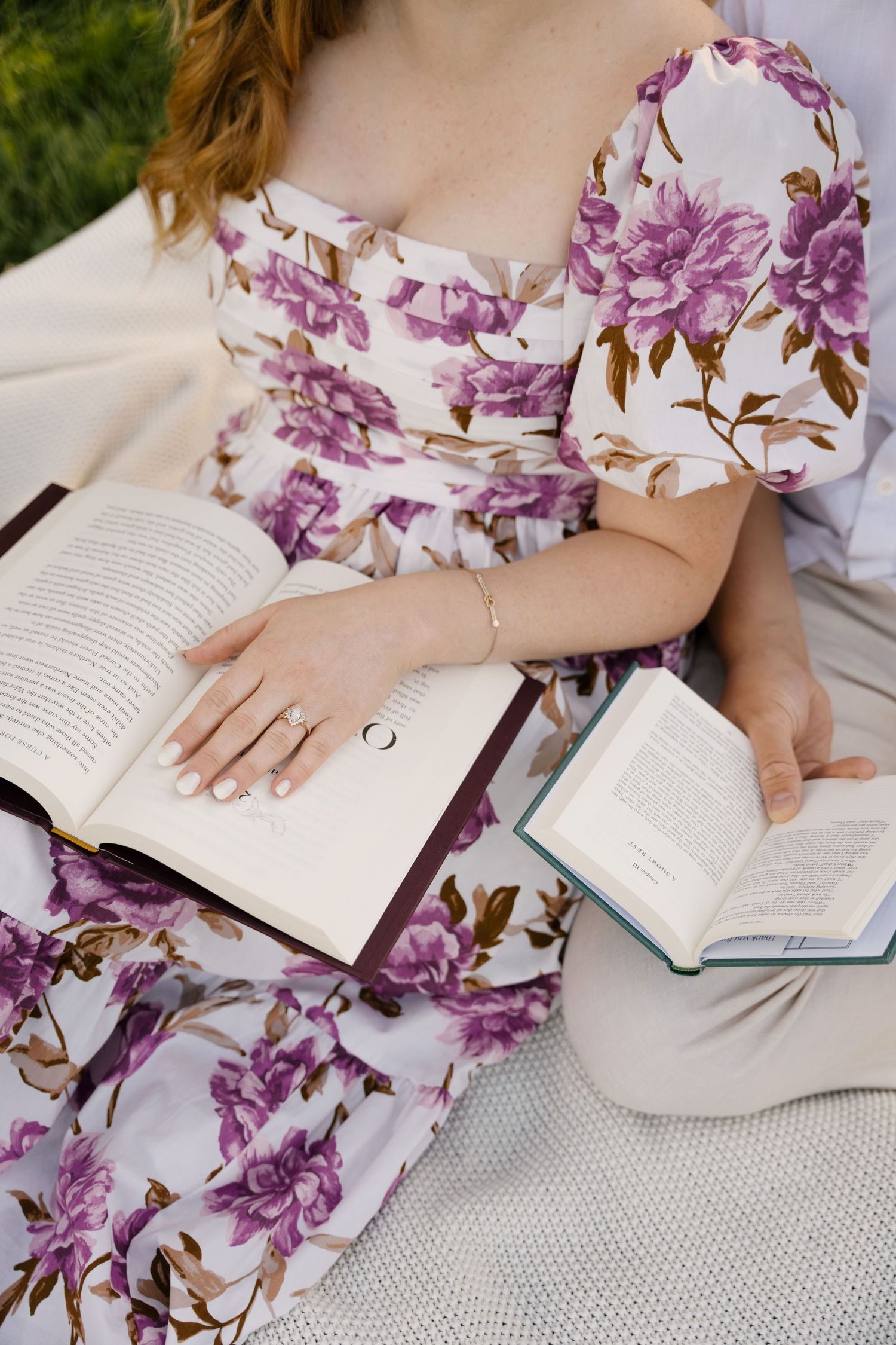 Woman in a floral dress sitting on a blanket with an engagement ring visible while holding books.