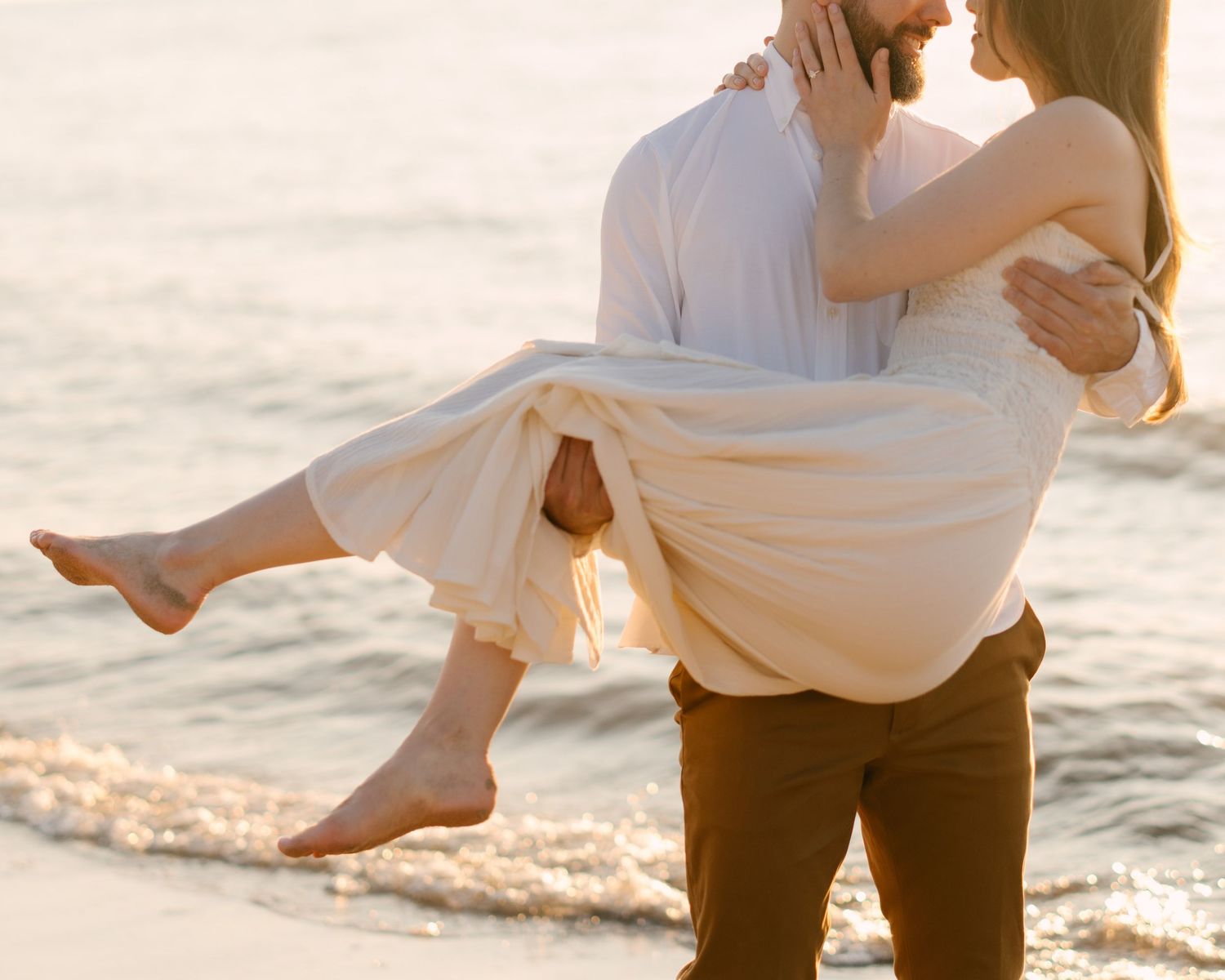 Man carrying woman in his arms on a sun-kissed beach at sunset, both dressed in light romantic attire.