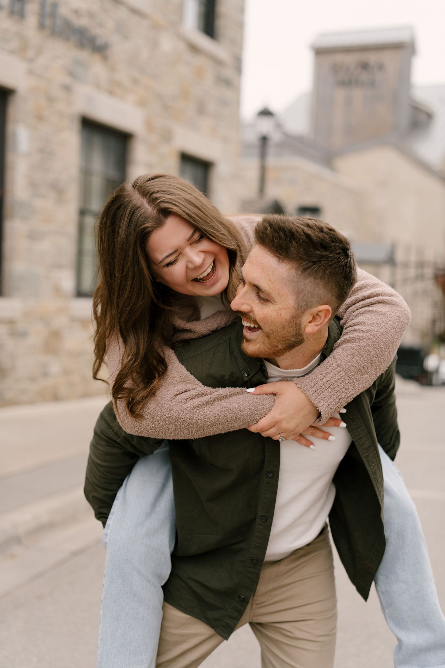 Laughing couple sharing a piggyback ride on a cobblestone street near stone buildings.