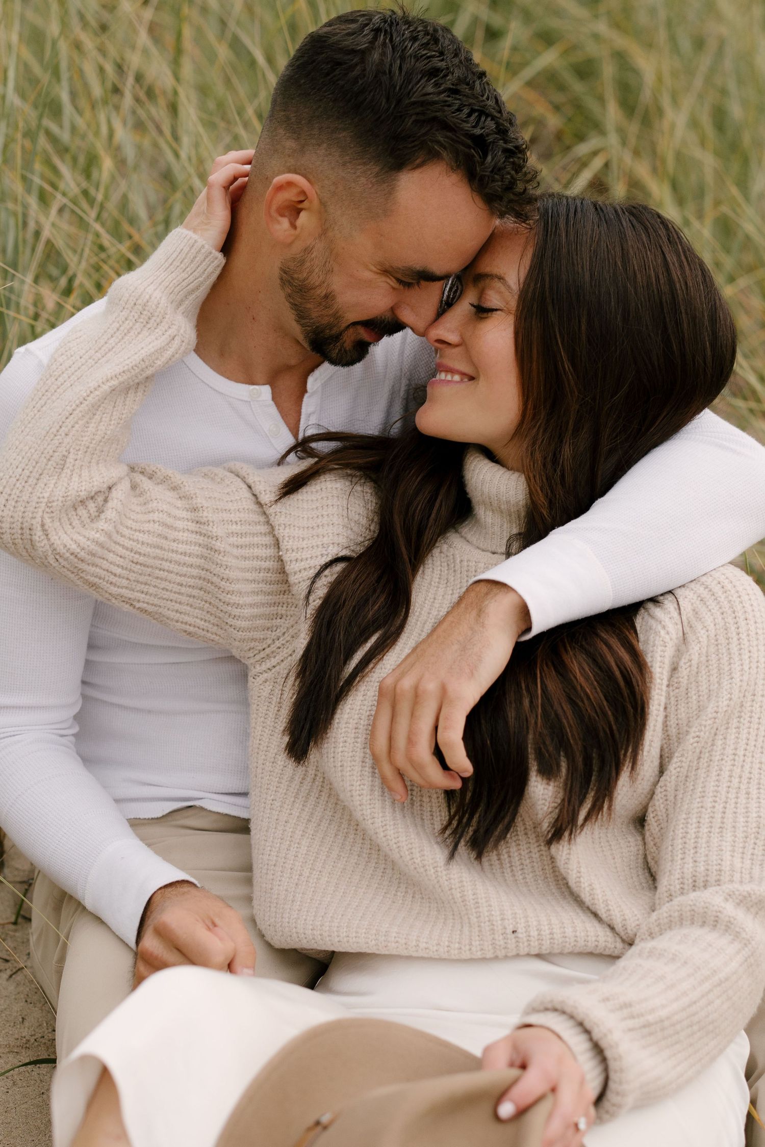 Romantic couple sitting close together in sand dunes, touching foreheads tenderly.