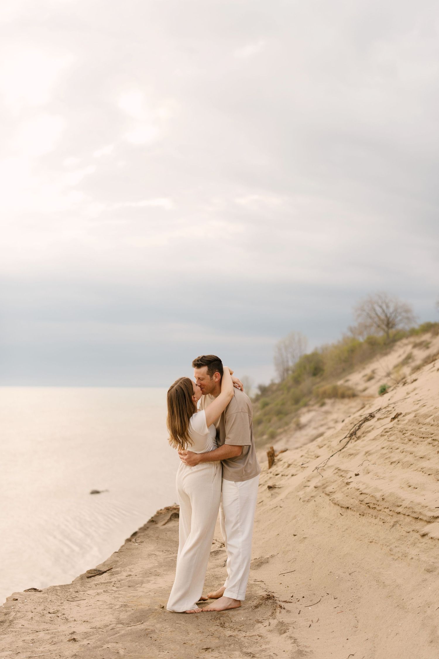 Couple sharing a kiss on a sandy beach dune with serene water in background.